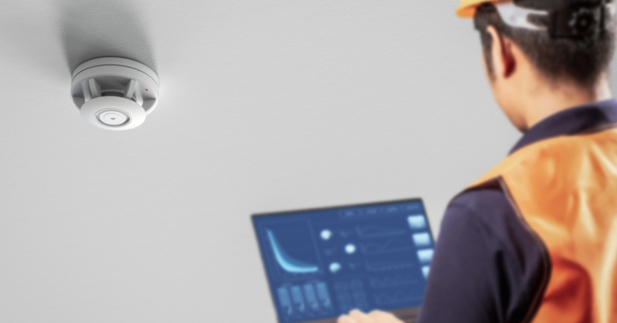An engineer programs a smoke detector with the help of a laptop. The engineer wears a hard hat and orange safety vest.