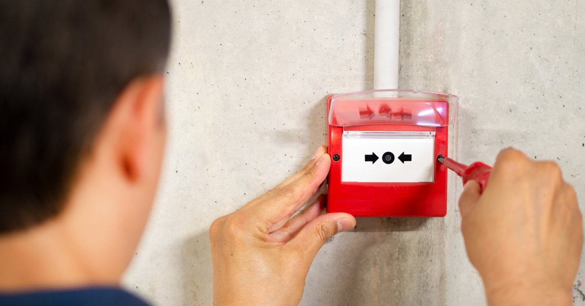 The back of an electrician installing a fire alarm system with a screwdriver. The system features two small arrows.