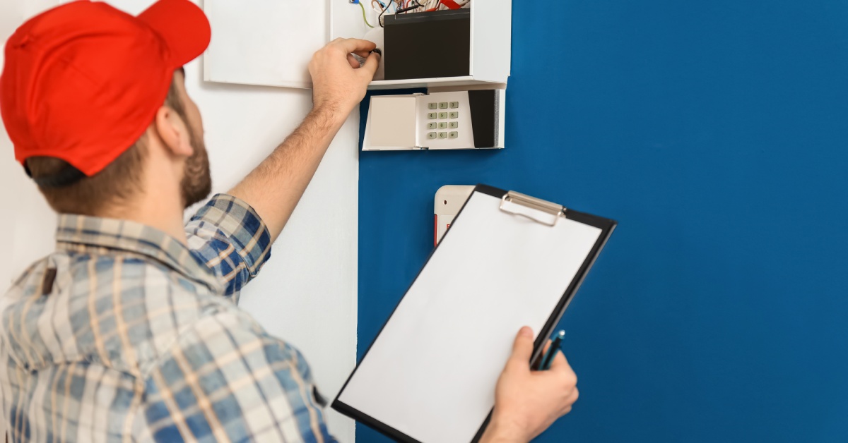 A man wearing a bright red cap looks at an alarm system on a wall. He holds a clipboard and a pen in his hand.