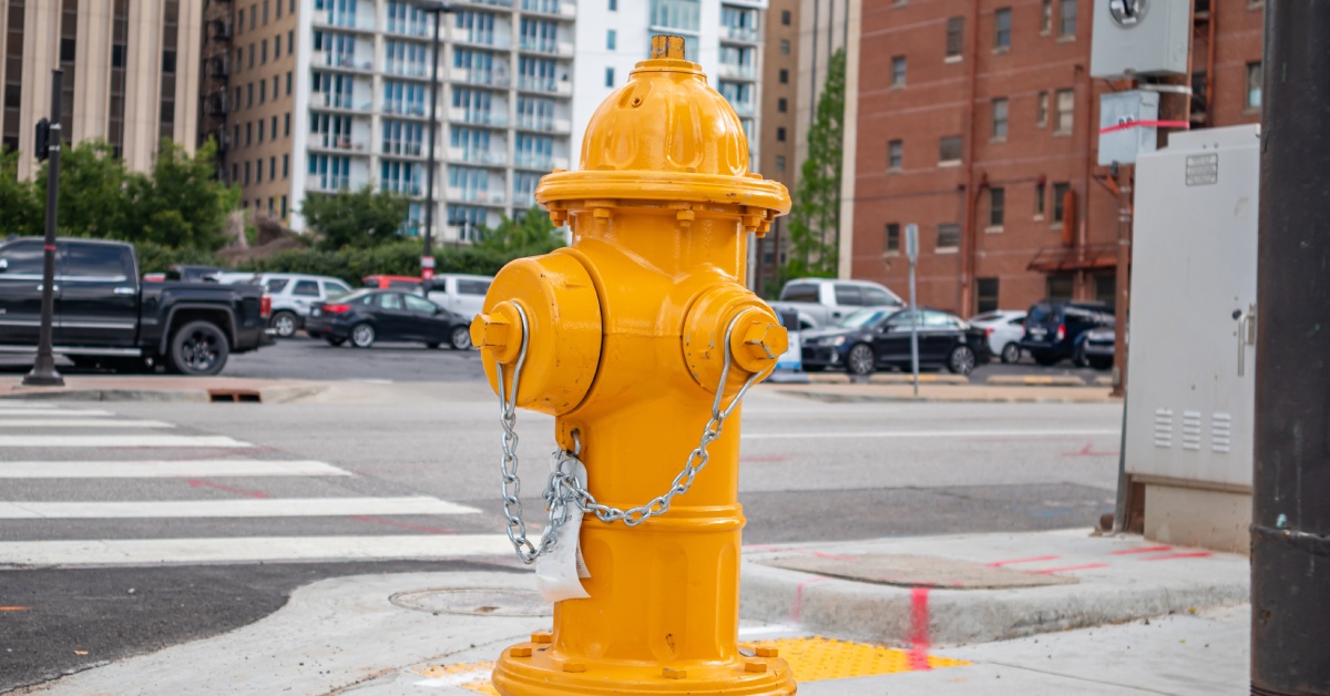 A yellow fire hydrant sits on a city street corner on a clear day. Several buildings stand in the background.