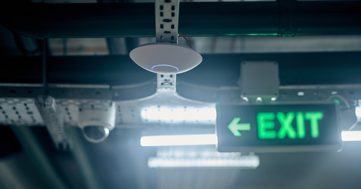 A circular fire alarm hangs from the ceiling. A sign reading "exit" and a security camera hang in the background behind it.
