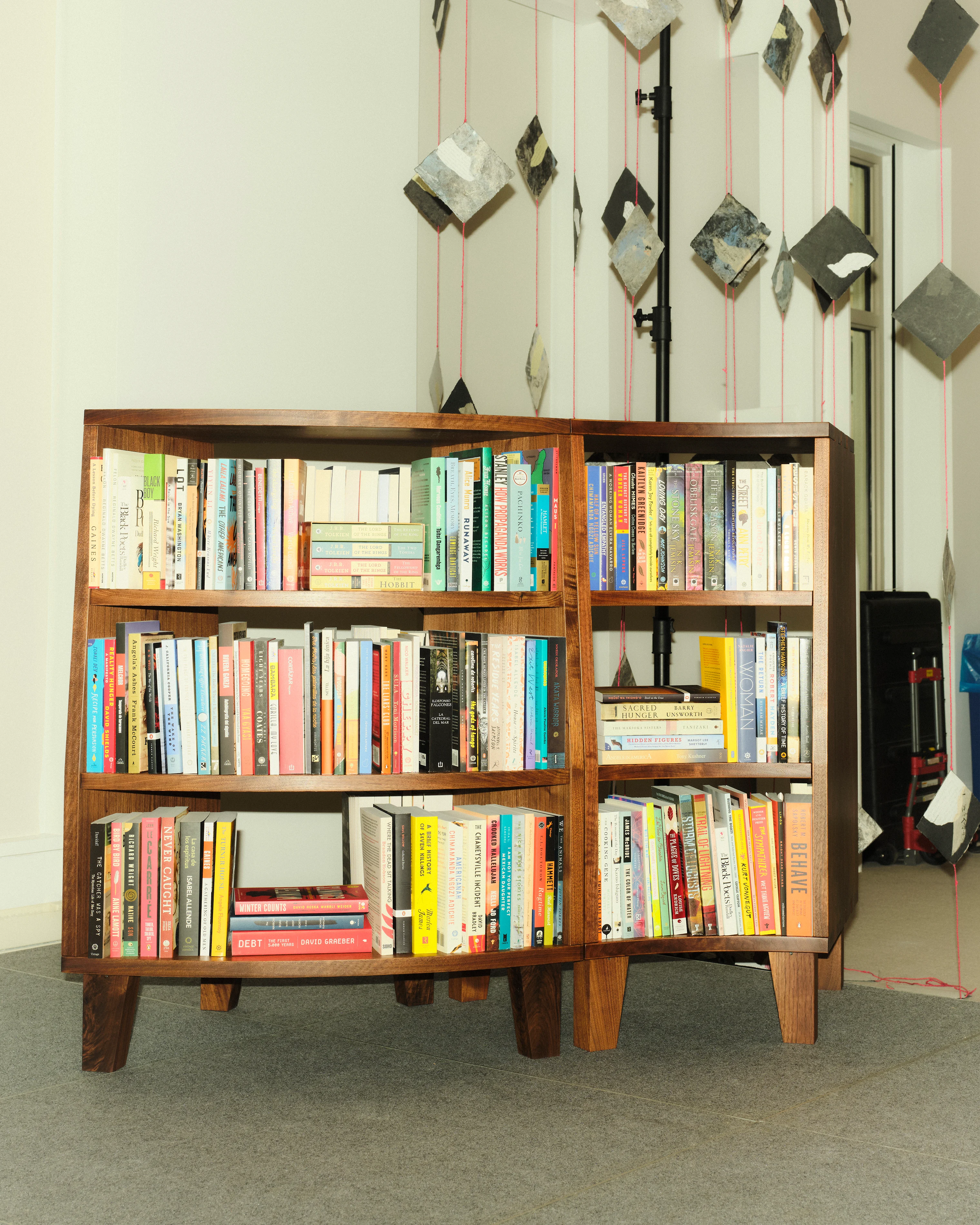 A wooden bookshelf filled with assorted books stands on a gray carpet. Hanging geometric decorations create a modern, artistic backdrop.