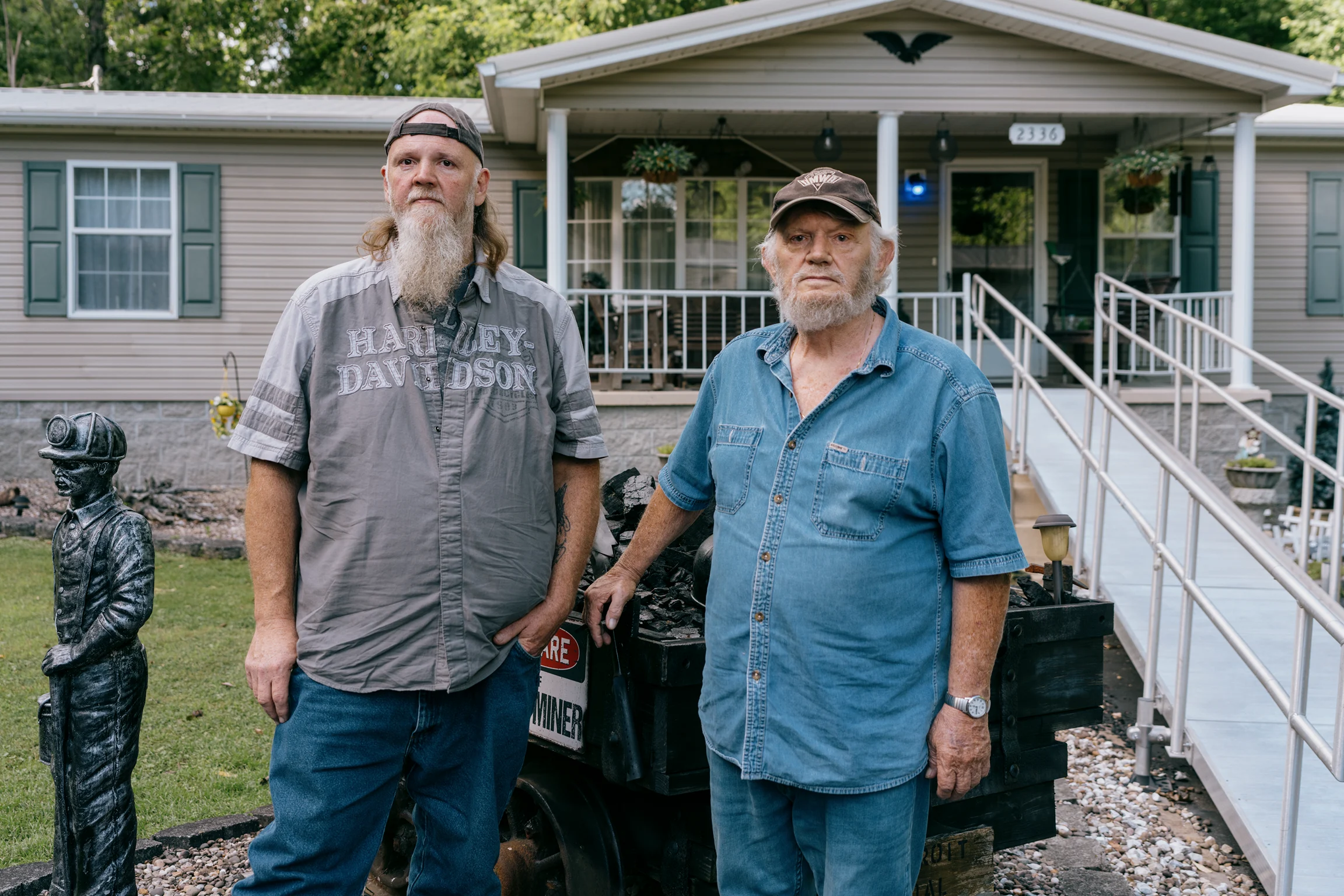 Two men stand beside a coal-filled mining cart and a miner statue in front of a single-story house with a covered porch and accessibility ramp.