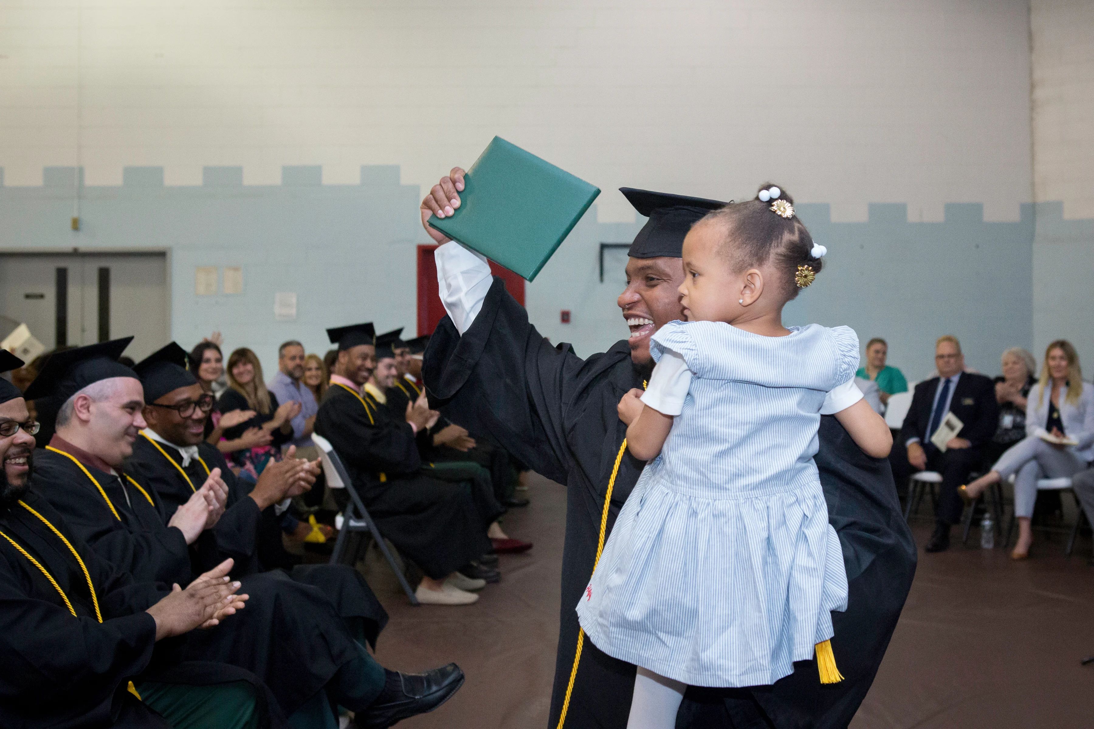 A smiling graduate wearing a cap and gown stands in a room before other graduates. The graduate is holding up a diploma and a child.