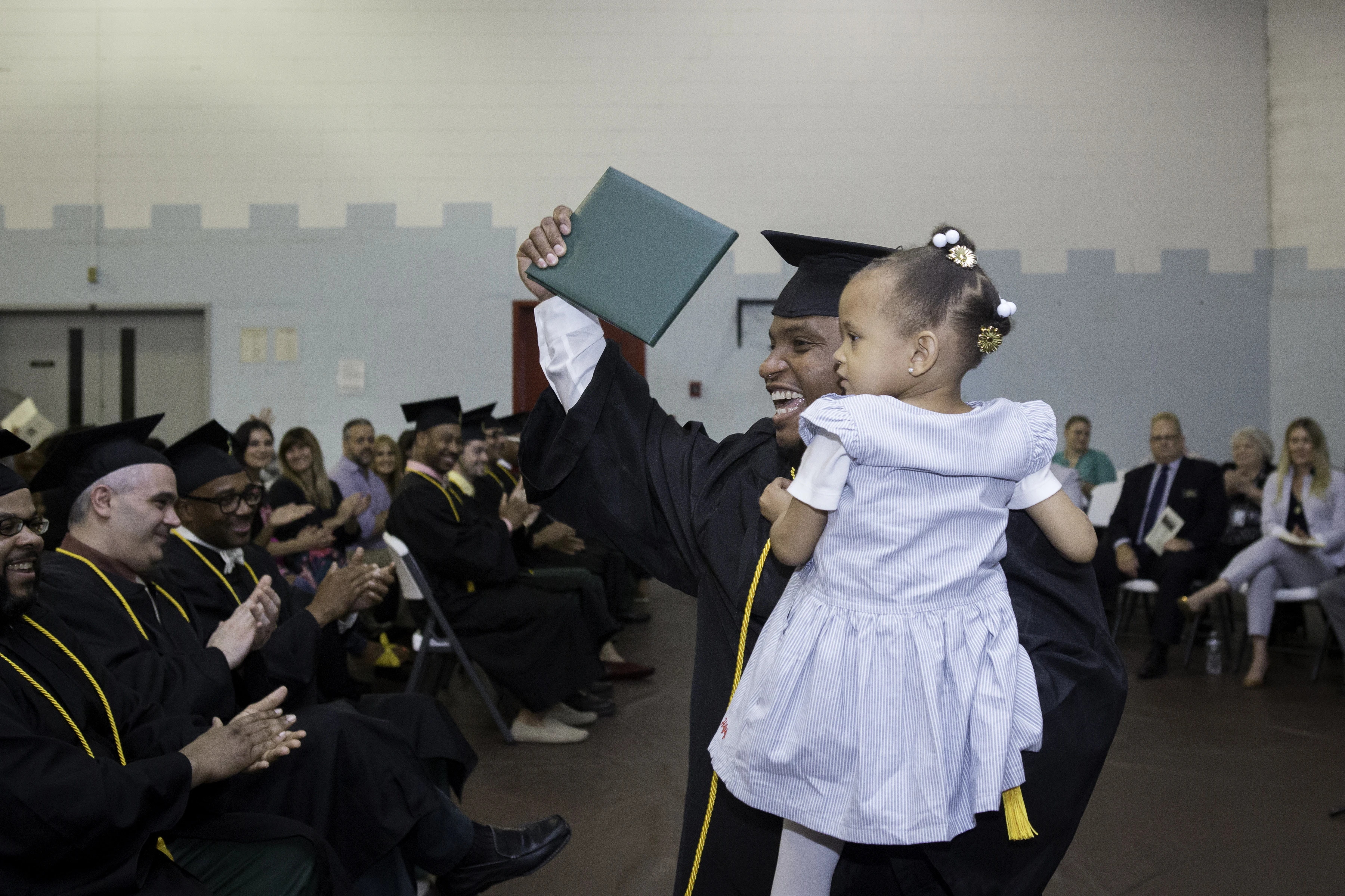 A smiling graduate wearing a cap and gown stands in a room before other graduates. The graduate is holding up a diploma and a child.