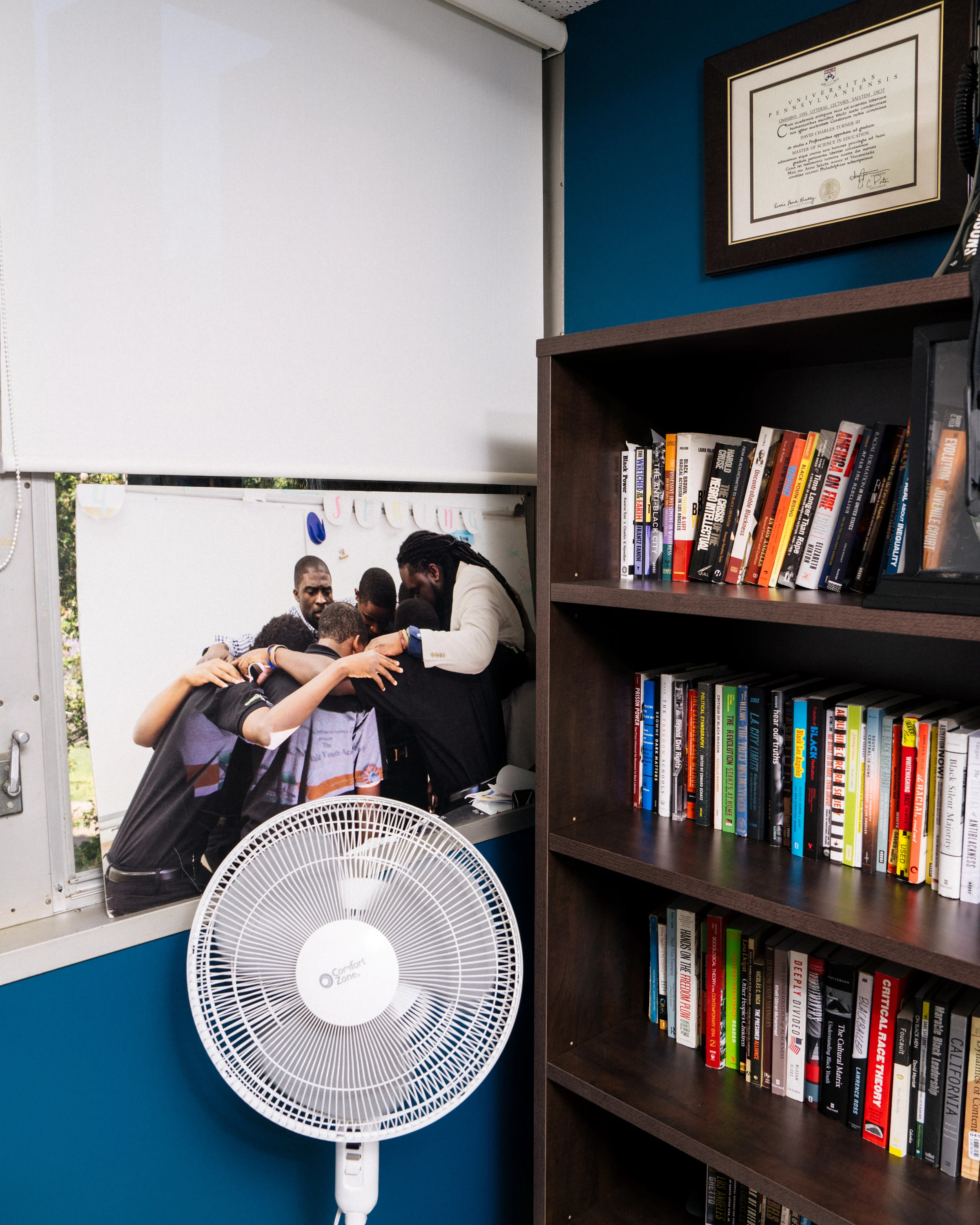 An office space with a bookshelf with books and a poster in the window of a group of people huddled together