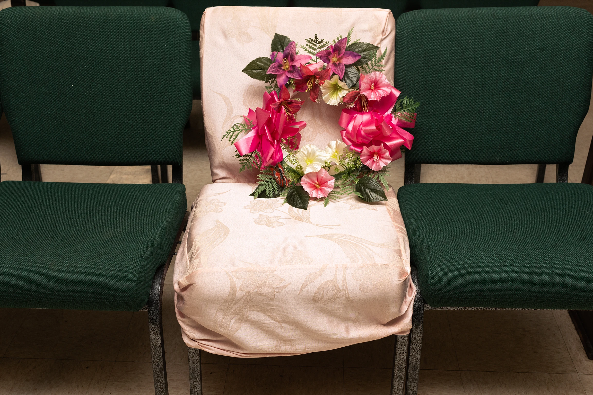 Three chairs set next to each other. The two on the ends have dark green fabric cushions. The middle is draped in a white cloth with a decorative floral wreath sitting propped up on it.