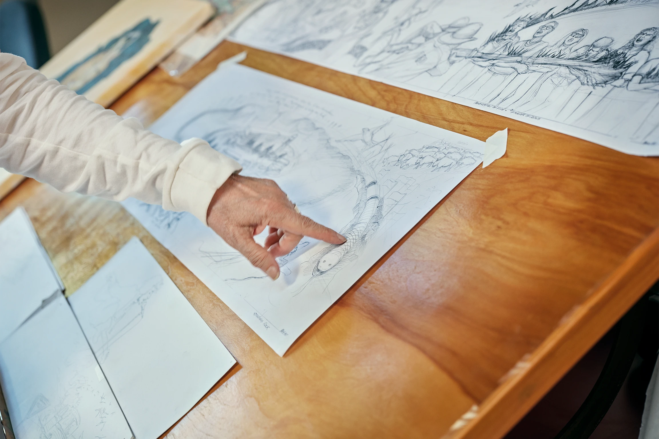 Multiple pieces of paper containing pencil sketches on a wooden table, with the artist Judy Baca's outstretched pointer finger touching one of them