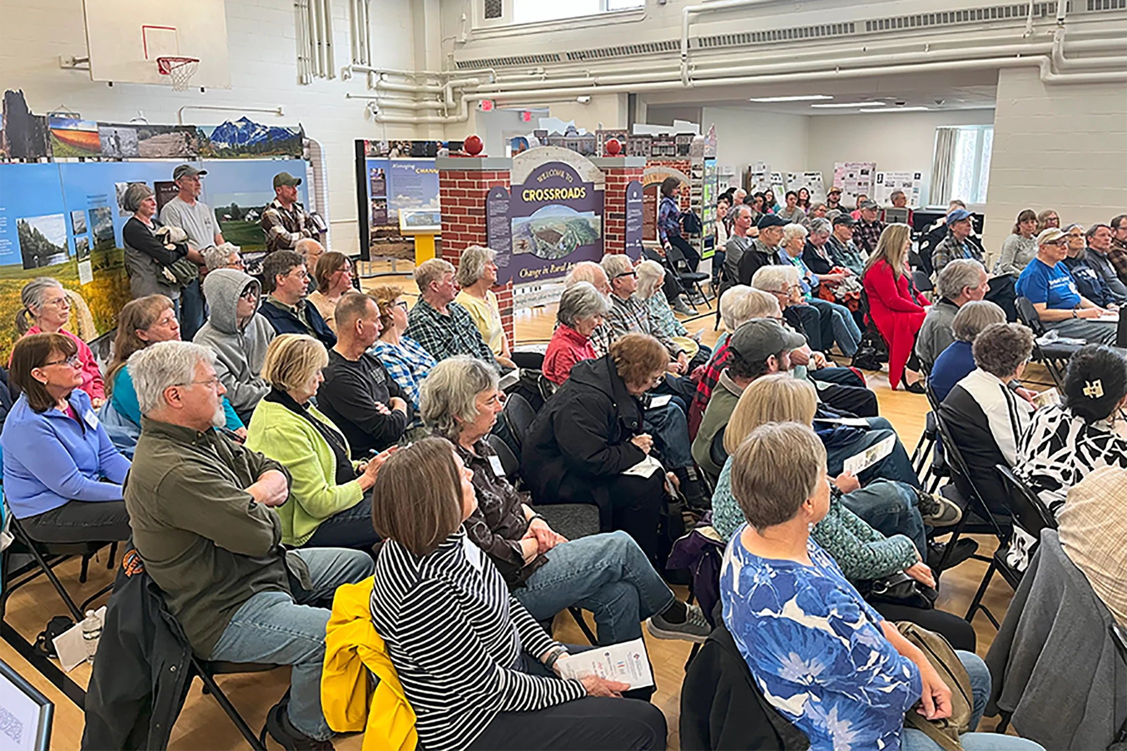 Un grupo grande de personas escucha una charla comunitaria en una sala similar a un gimnasio con paneles de exhibición sobre la historia local y el cambio social.