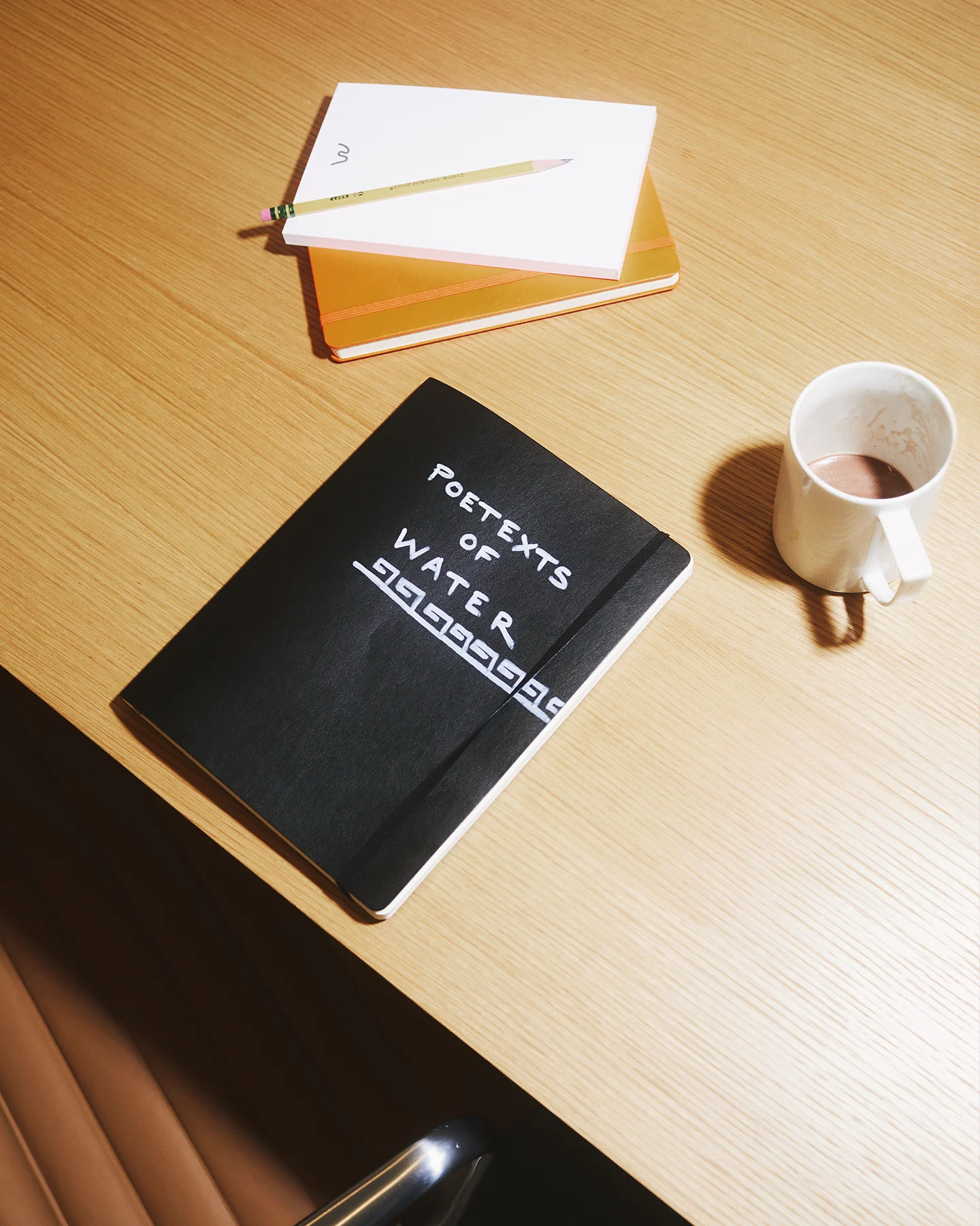A notebook with a black cover, a mug, and a pad of paper are sitting on a table. The text on the cover of the black notebook reads, "Poetexts of Water."