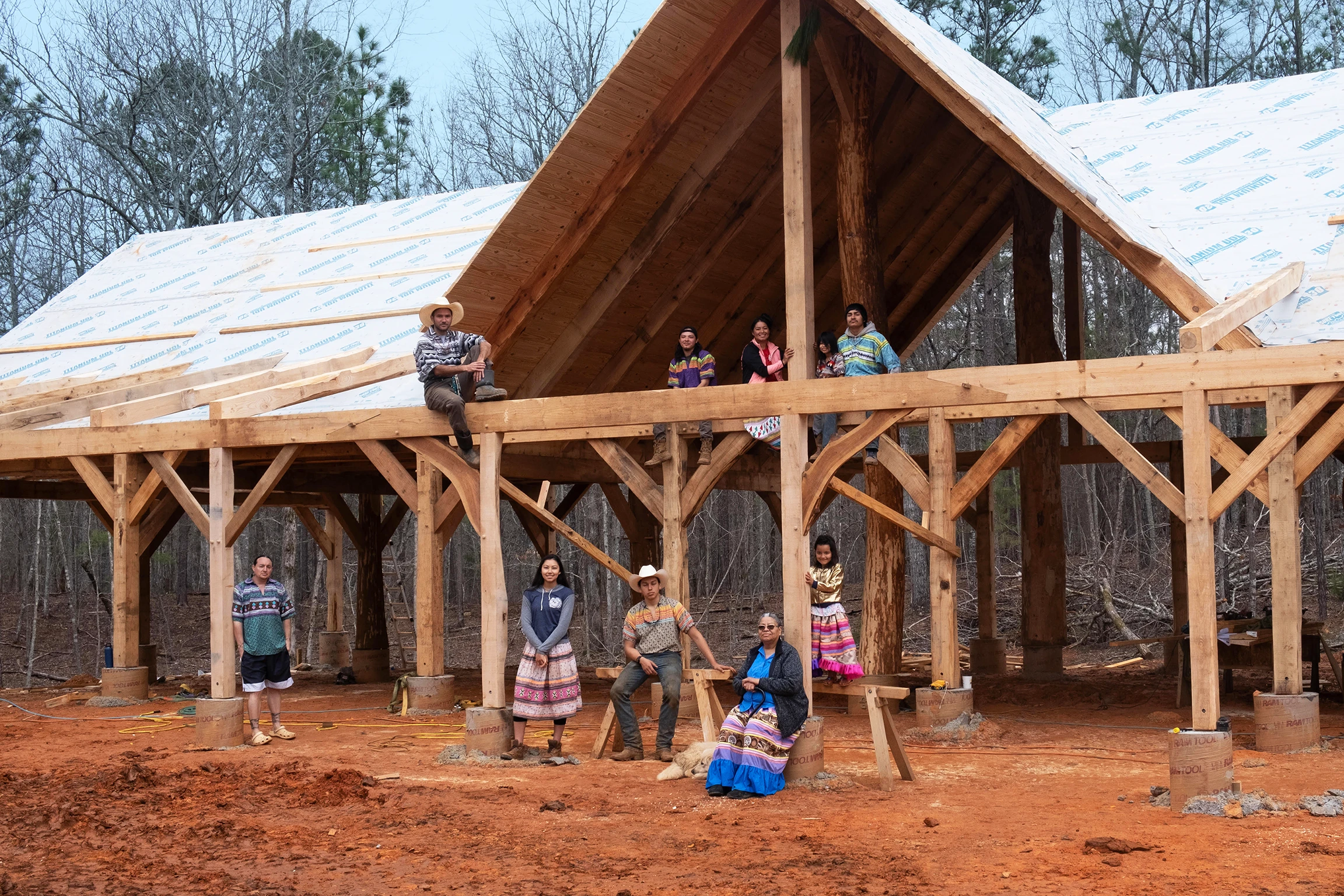 A group of Indigenous Maskoke people stand in an open frame of a building that is set against a woods.