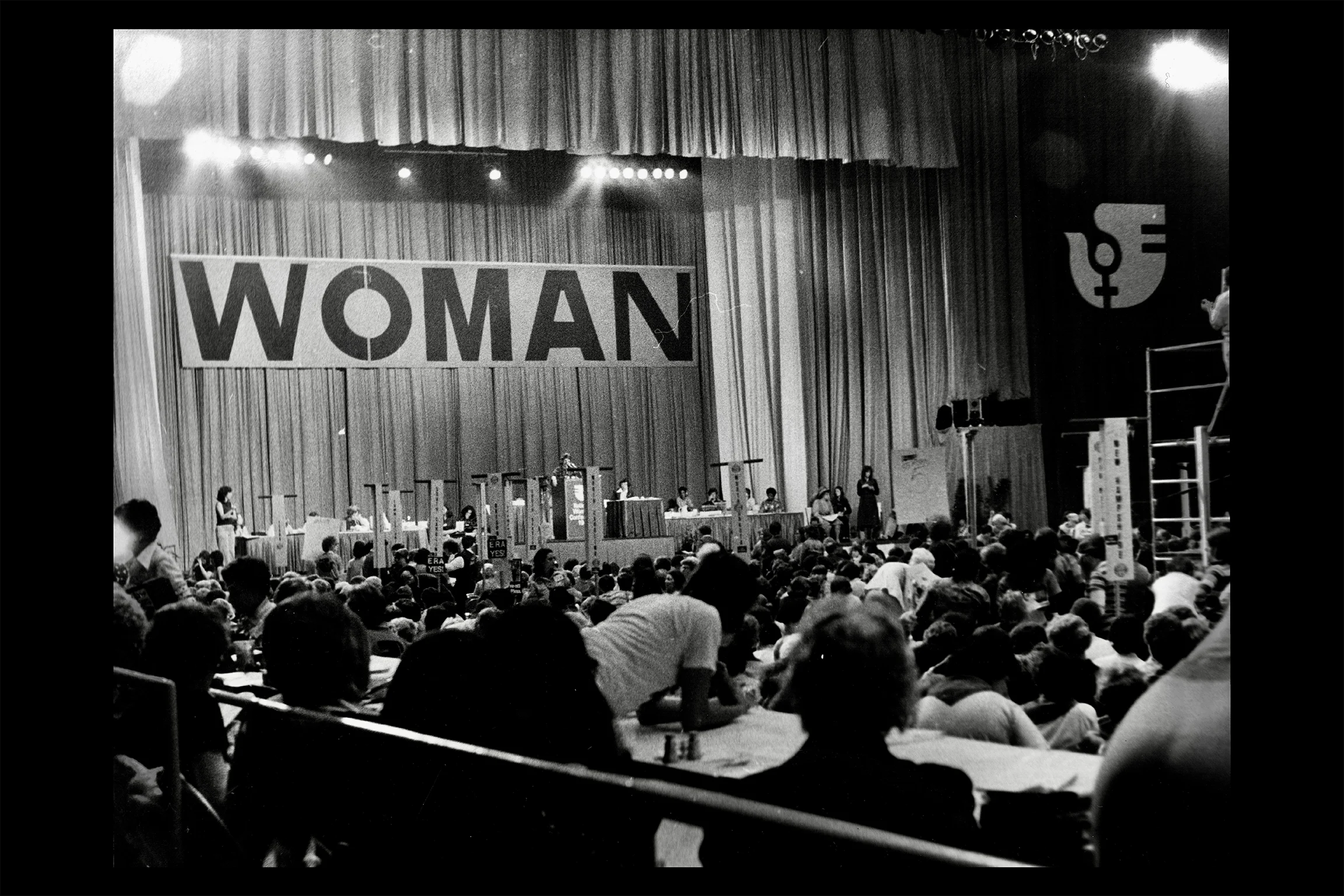 Black-and-white photograph of a large women’s conference or convention, with a crowded auditorium facing a stage beneath a large banner reading “WOMAN” and a feminist symbol displayed to the side.
