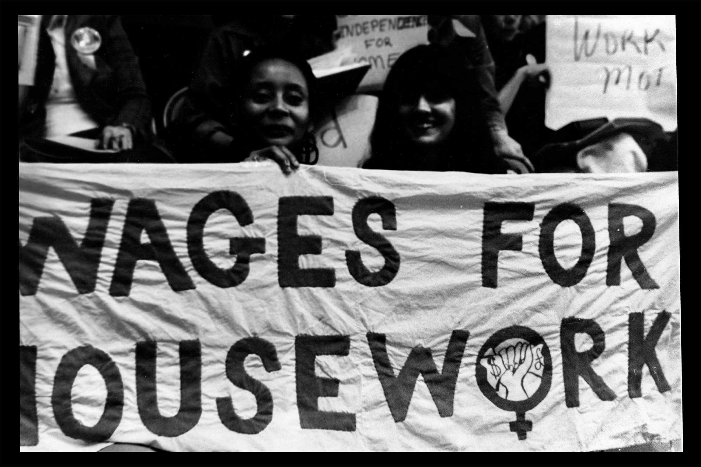 Black-and-white photograph of protesters holding a large banner reading “WAGES FOR HOUSEWORK,” with additional handmade signs visible in the background.