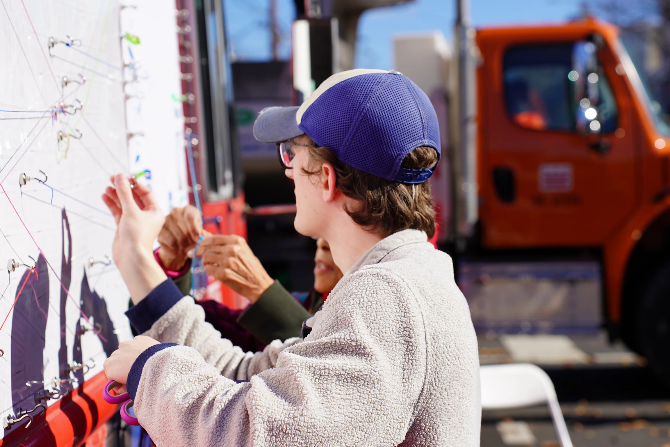 A male wearing glasses and a blue baseball cap ties string to hooks that are on the side of a large truck