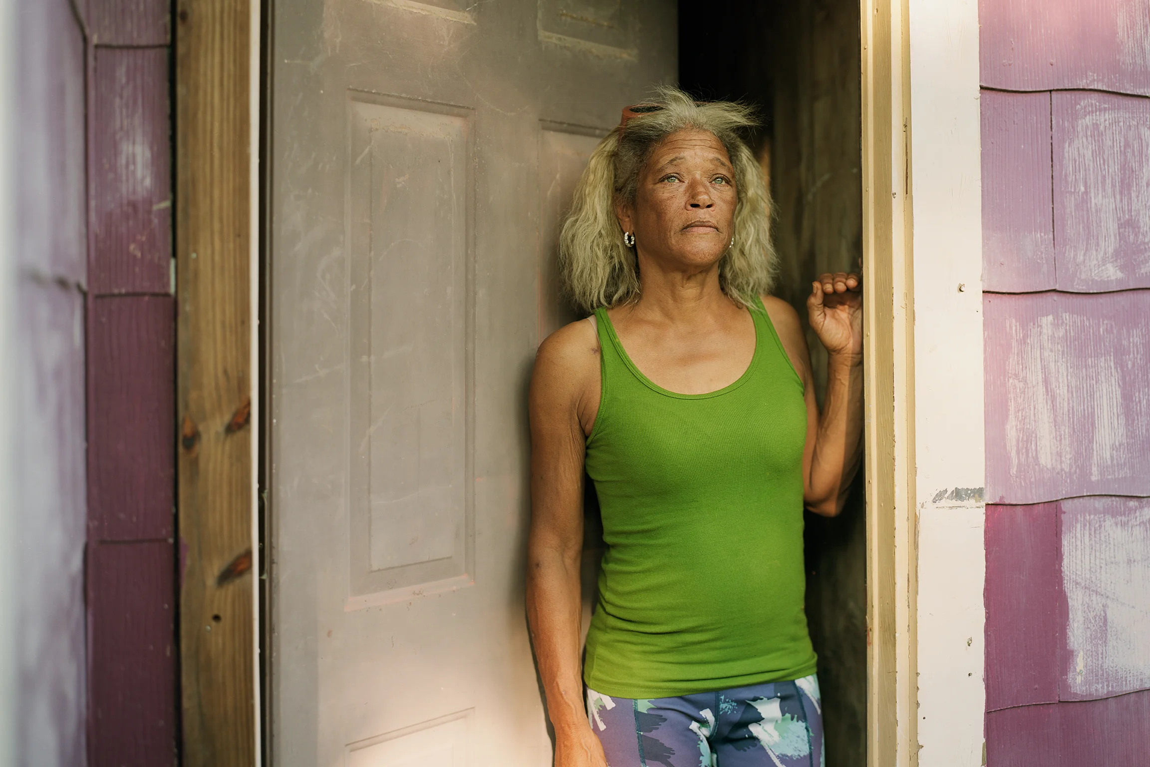 Deidra Dunn-Pleasant wears a tank top and patterned leggings as she leans against the door frame of a paneled house.