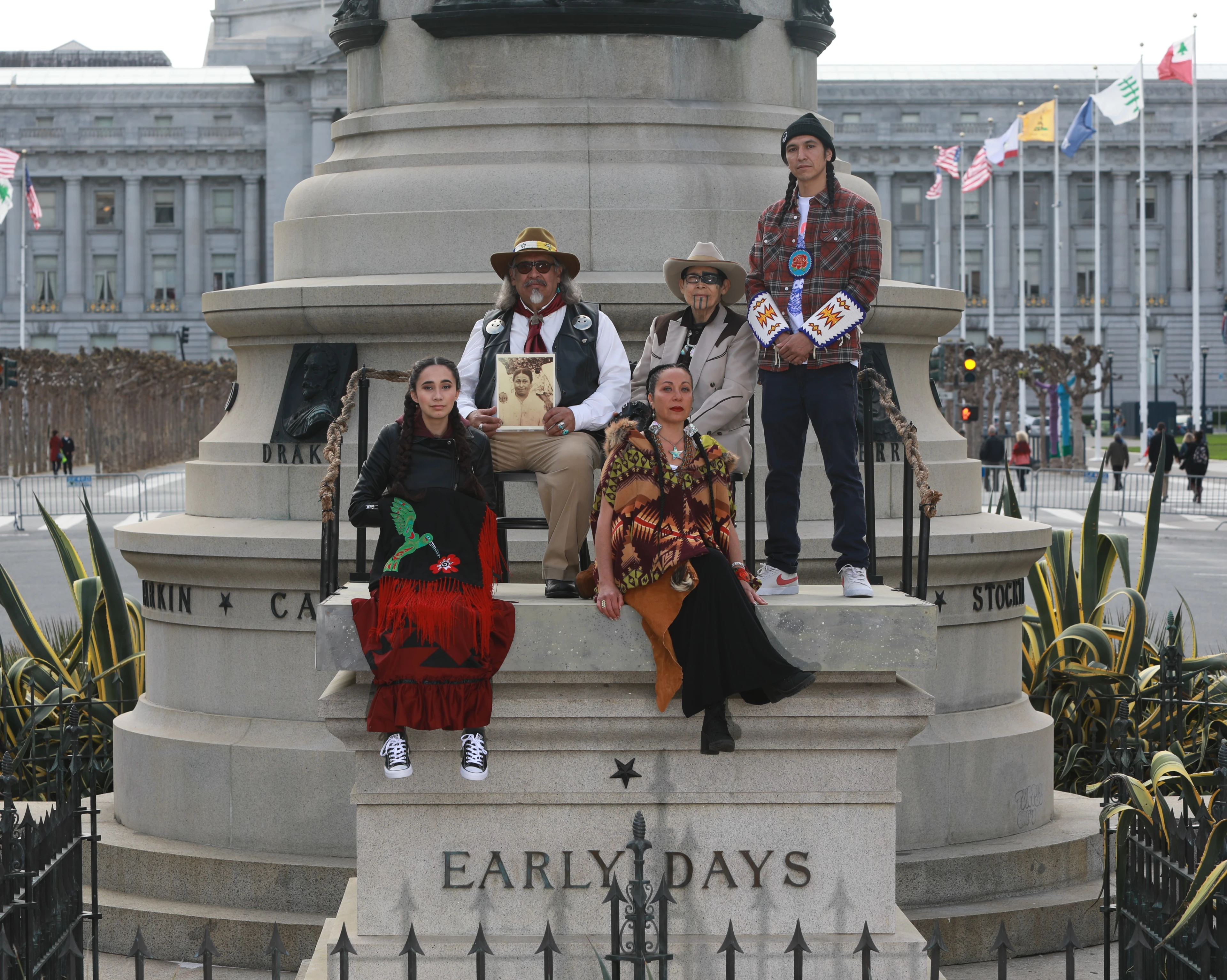 Five Indigenous artists stand and sit in front of a monument in San Francisco