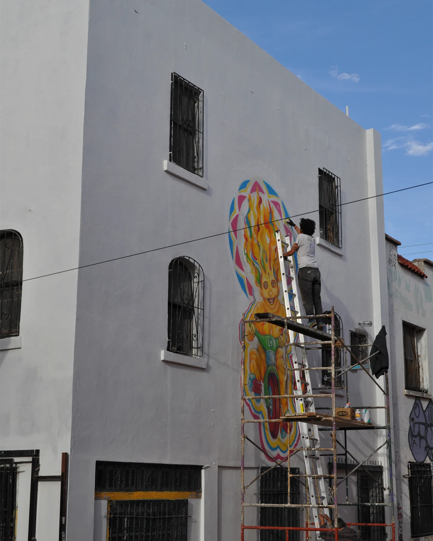 An artist on scaffolding painting a mural on the exterior of a white concrete building
