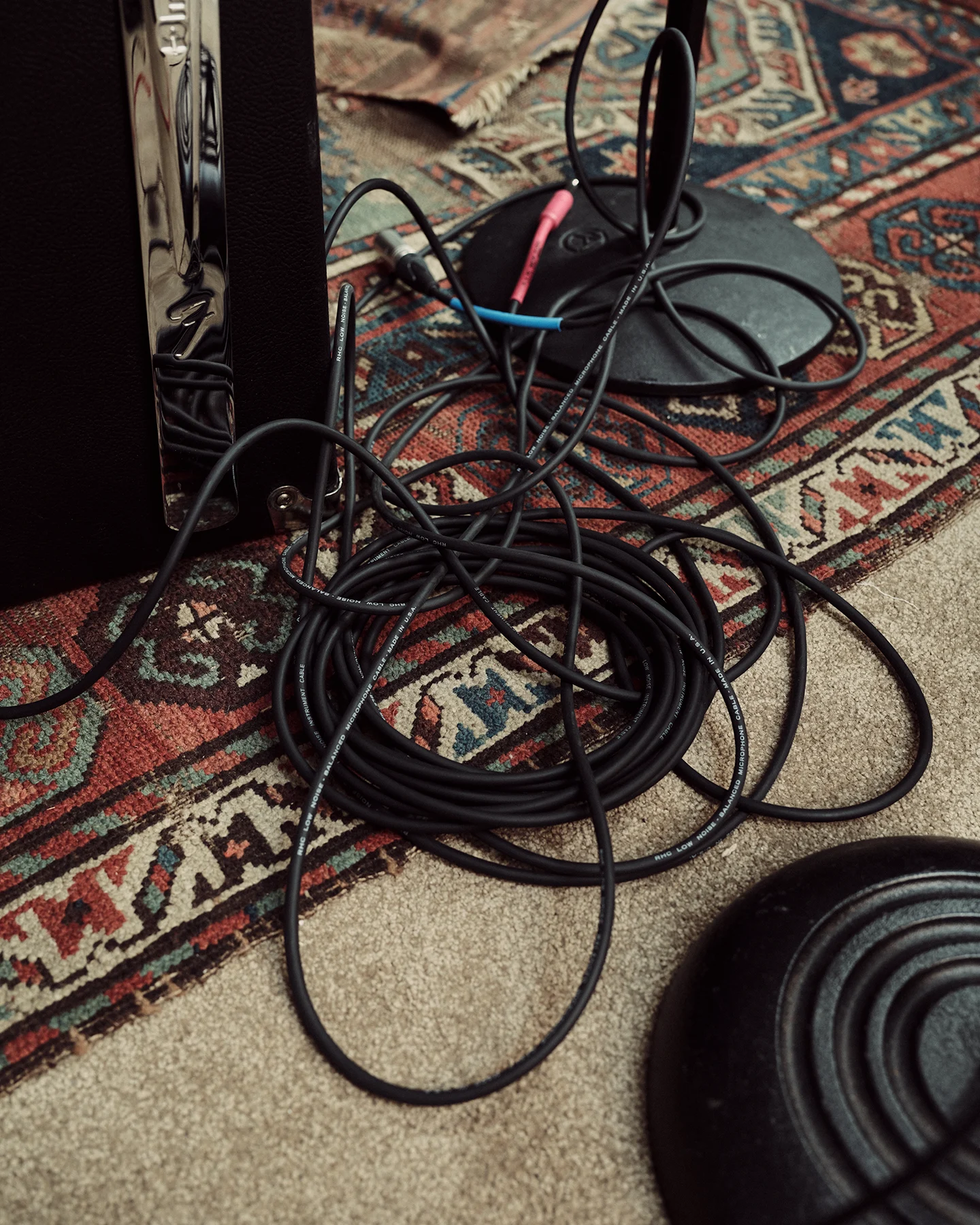 Close-up of coiled audio cables on a patterned rug beside the base of a microphone stand and an amplifier. The image highlights the behind-the-scenes equipment used in a music studio.