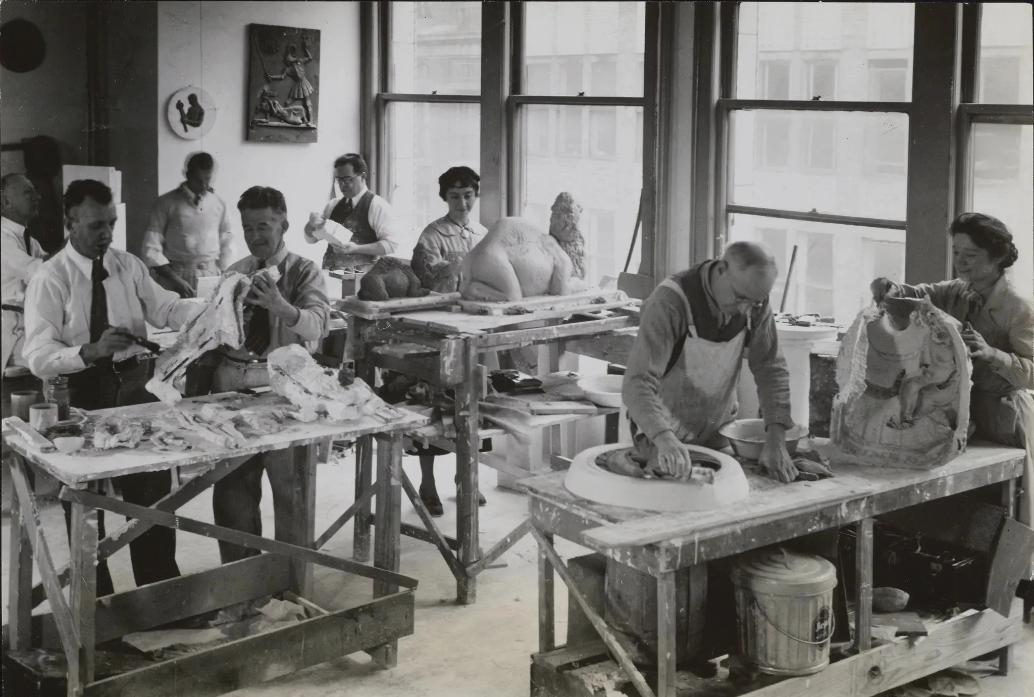 Artists work in pairs around small paint-stained benches in a messy studio. In black and white, their hair and clothes indicate that this photo was taken in the early 1900s. Artists work on various stages of sculptures. One artist paints a statue of a camel.
