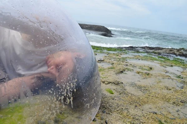 A close-up shot on a mossy beach shows a person in the foreground laying on their side in a clear bubble.