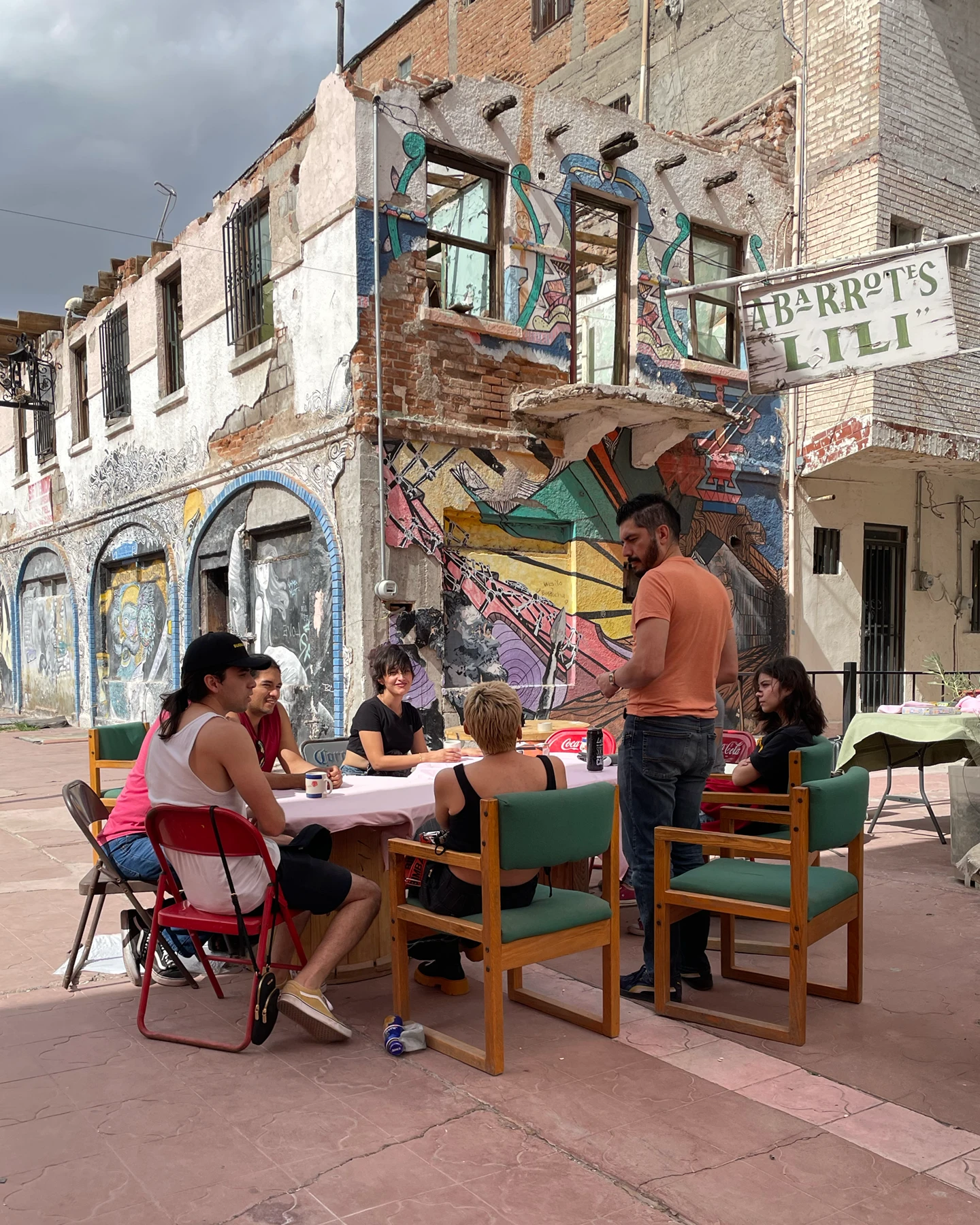 A group of people sit and stand around a table covered in a white cloth outside on a sidewalk along a graffiti covered wall