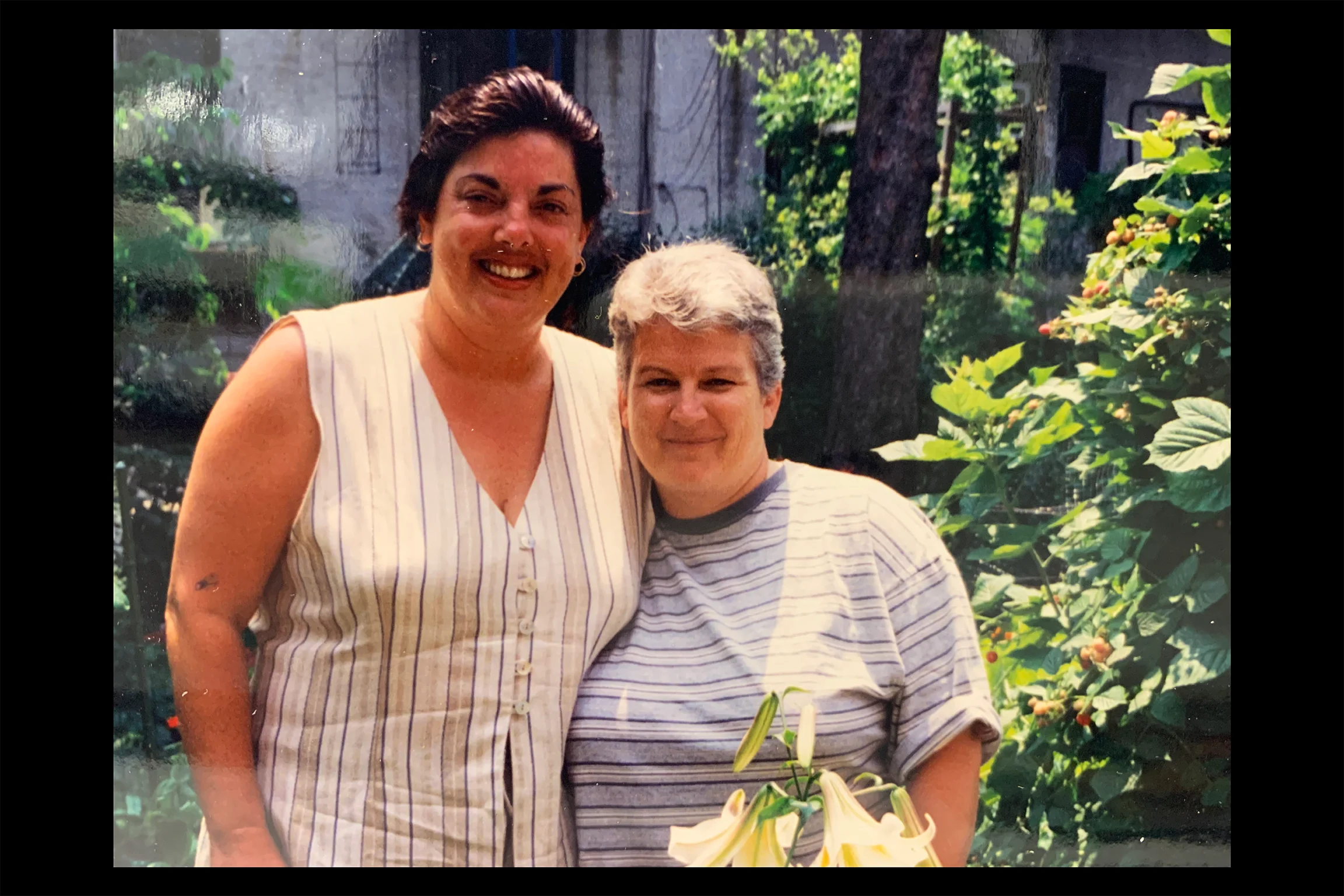 Color photograph of two women standing together outdoors in a garden, smiling at the camera beside flowering plants.