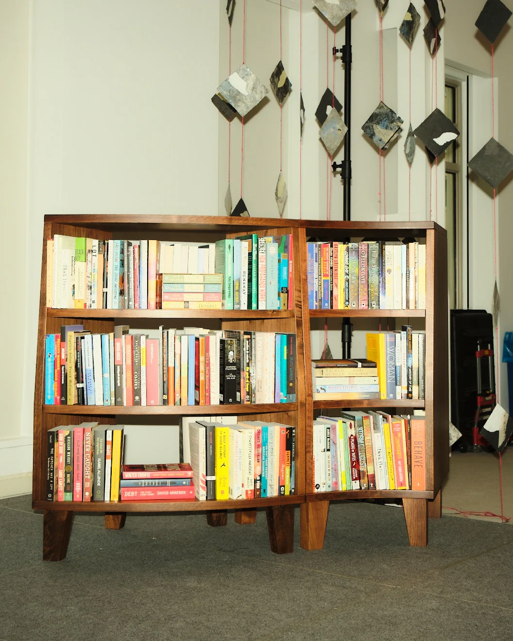 A mid-century-style wooden bookshelf filled with a variety of books stands on a carpeted floor. The books are arranged across three shelves, and an abstract hanging installation of diamond-shaped pieces suspended on red strings decorates the background.