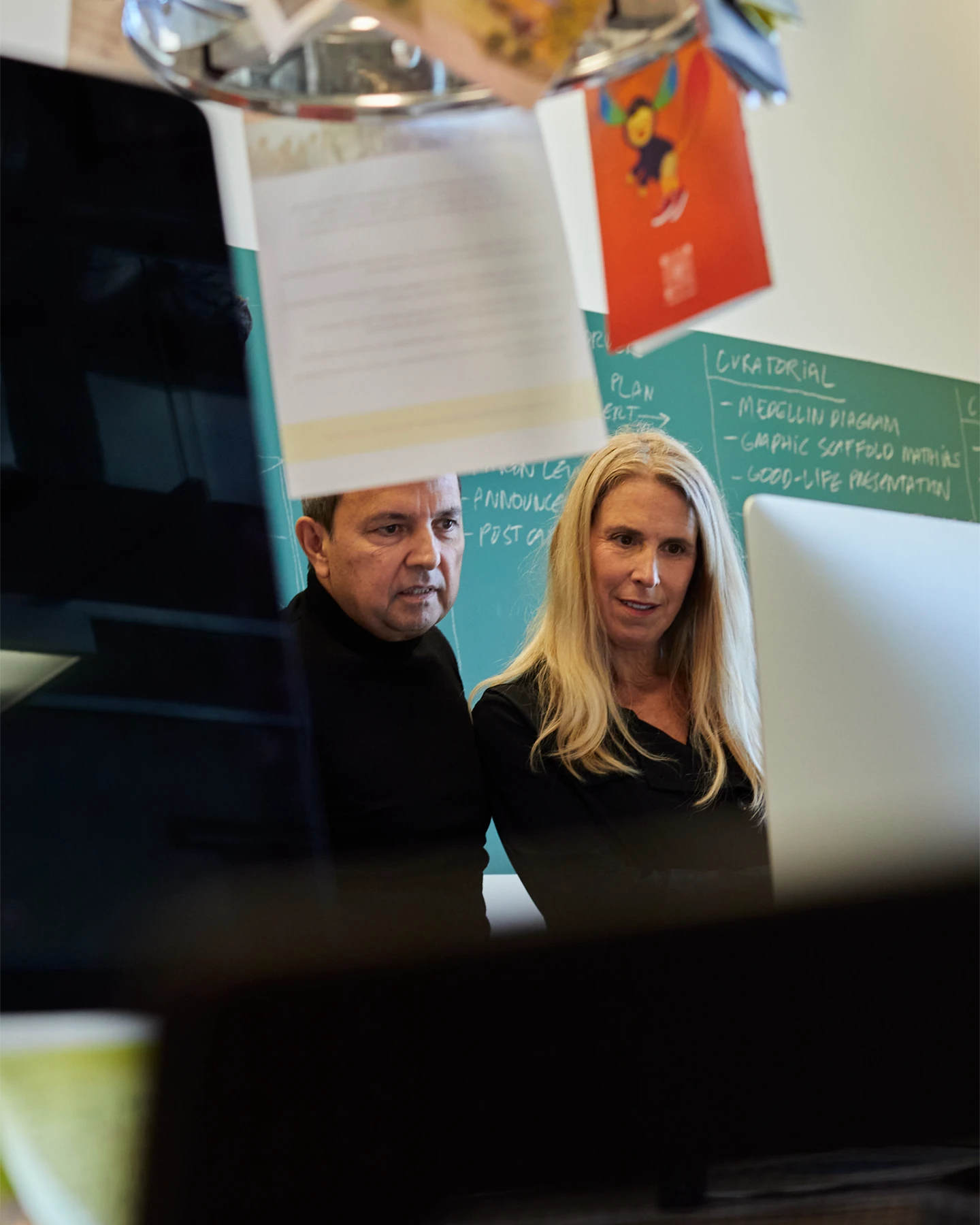 A man and a woman standing side by side looking at a computer monitor with a chalkboard behind them 