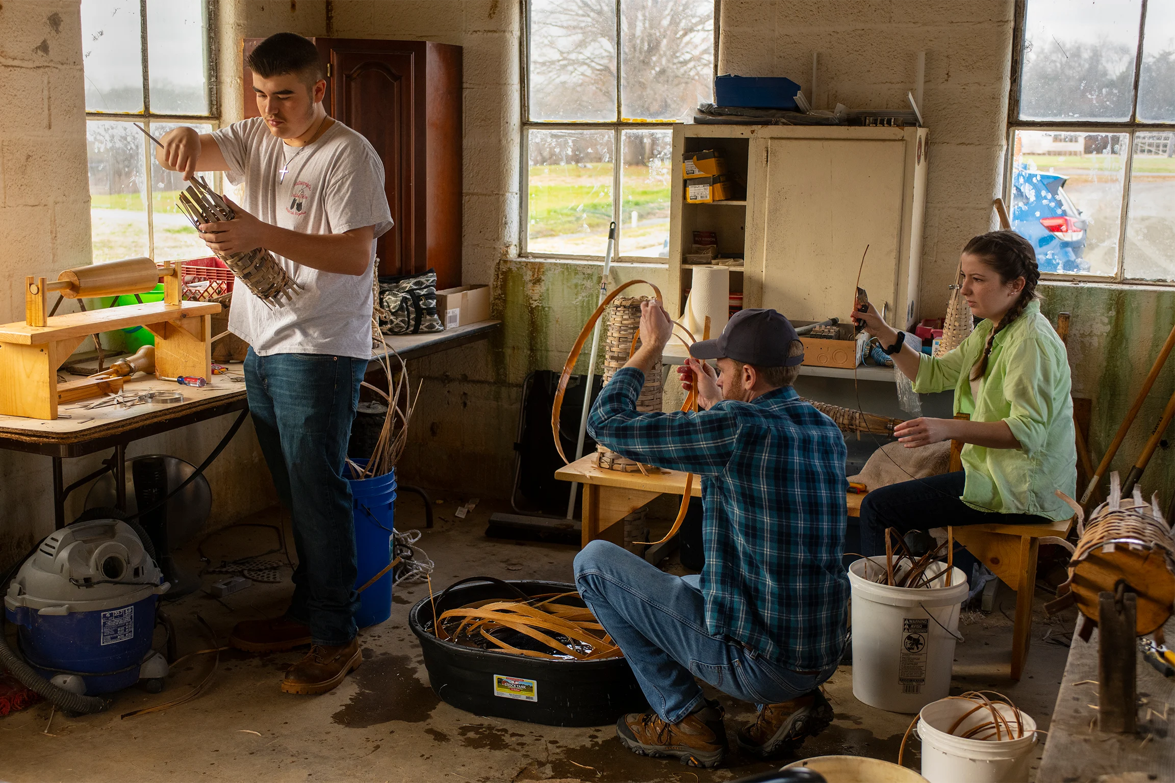 Three people weave eel pots in a bright workshop filled with tools, buckets, and materials, with windows showing a rural view outside.