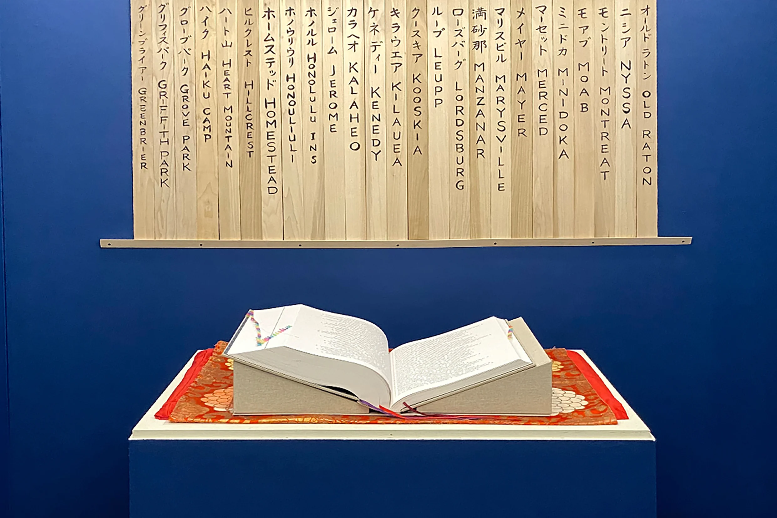 "Ireichō book" sits on a blue pedestal. A series of vertical slats with names of formerly incarcerated people of Japanese descent hangs on the wall.