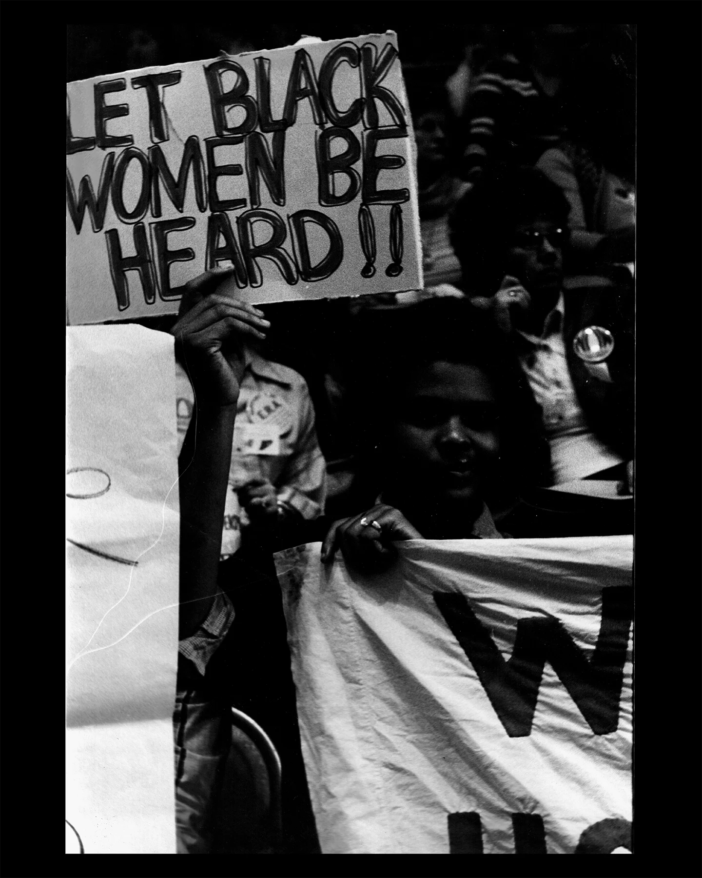 Black-and-white photograph of protesters at a rally, including a person holding a sign reading “LET BLACK WOMEN BE HEARD!!” beside a partially visible “Wages for Housework” banner.