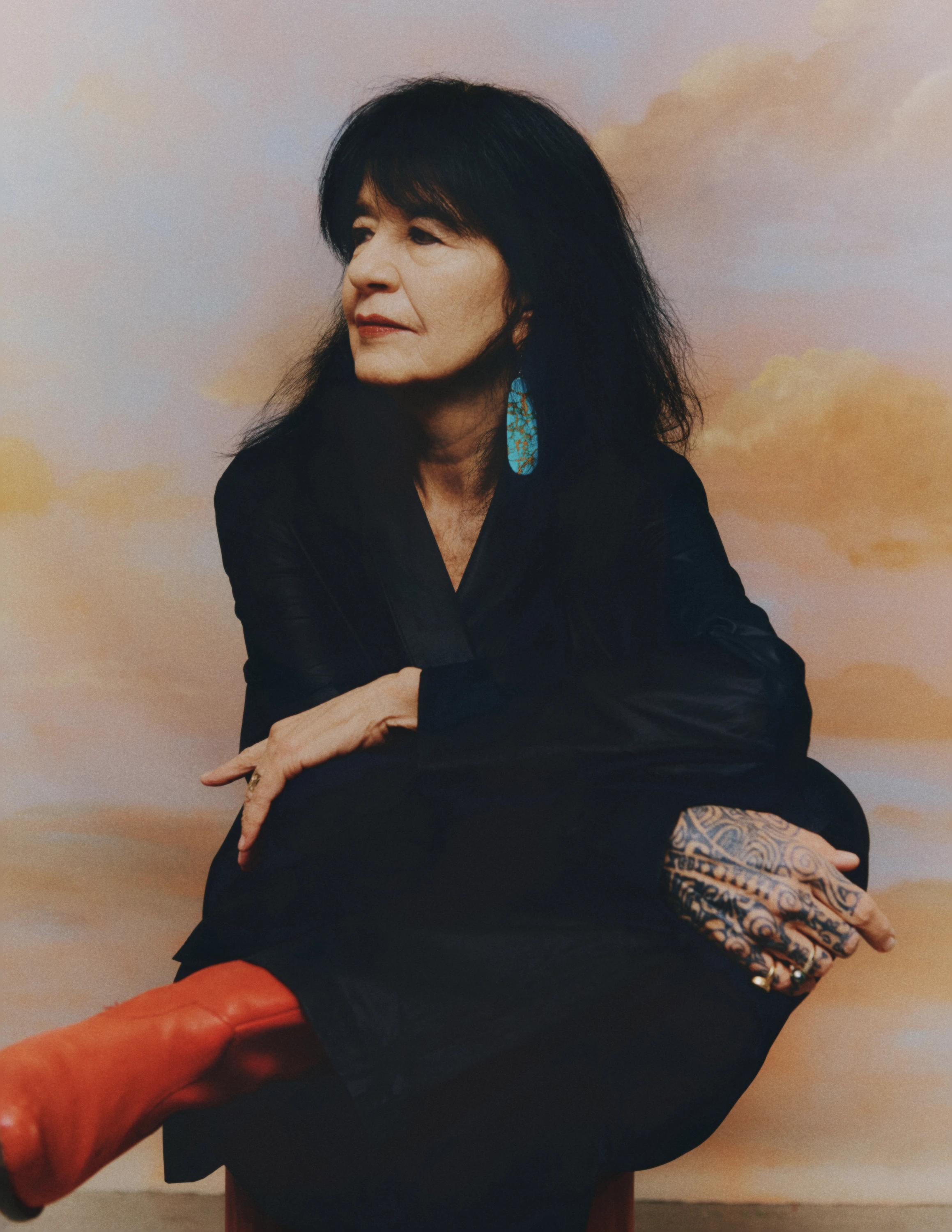 A portrait of Joy Harjo, a Native American woman wearing all black, with tattoos on her right hand and wearing red boots