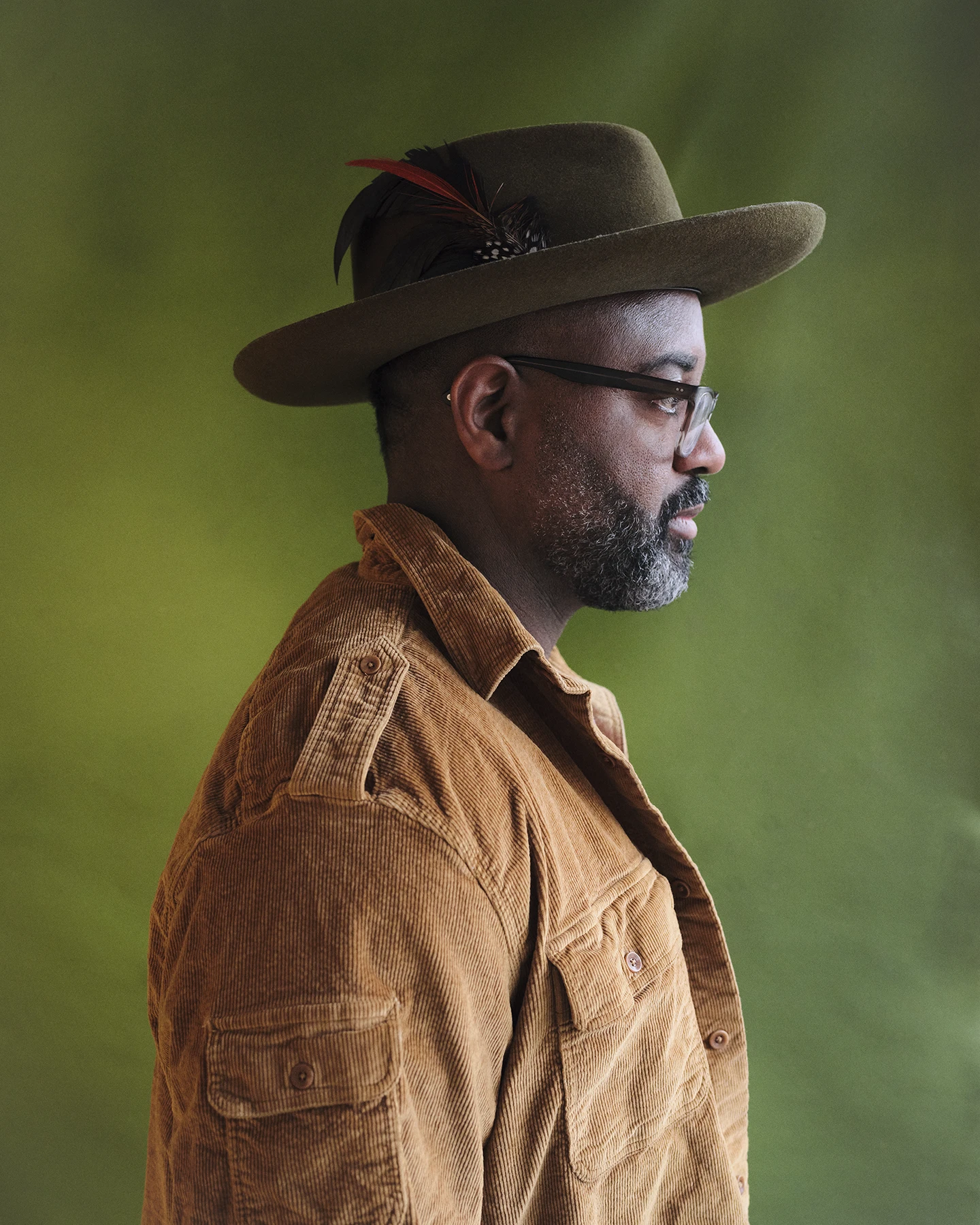 A side profile of a Black poet who is wearing glasses and a hat with a feather hat pin.