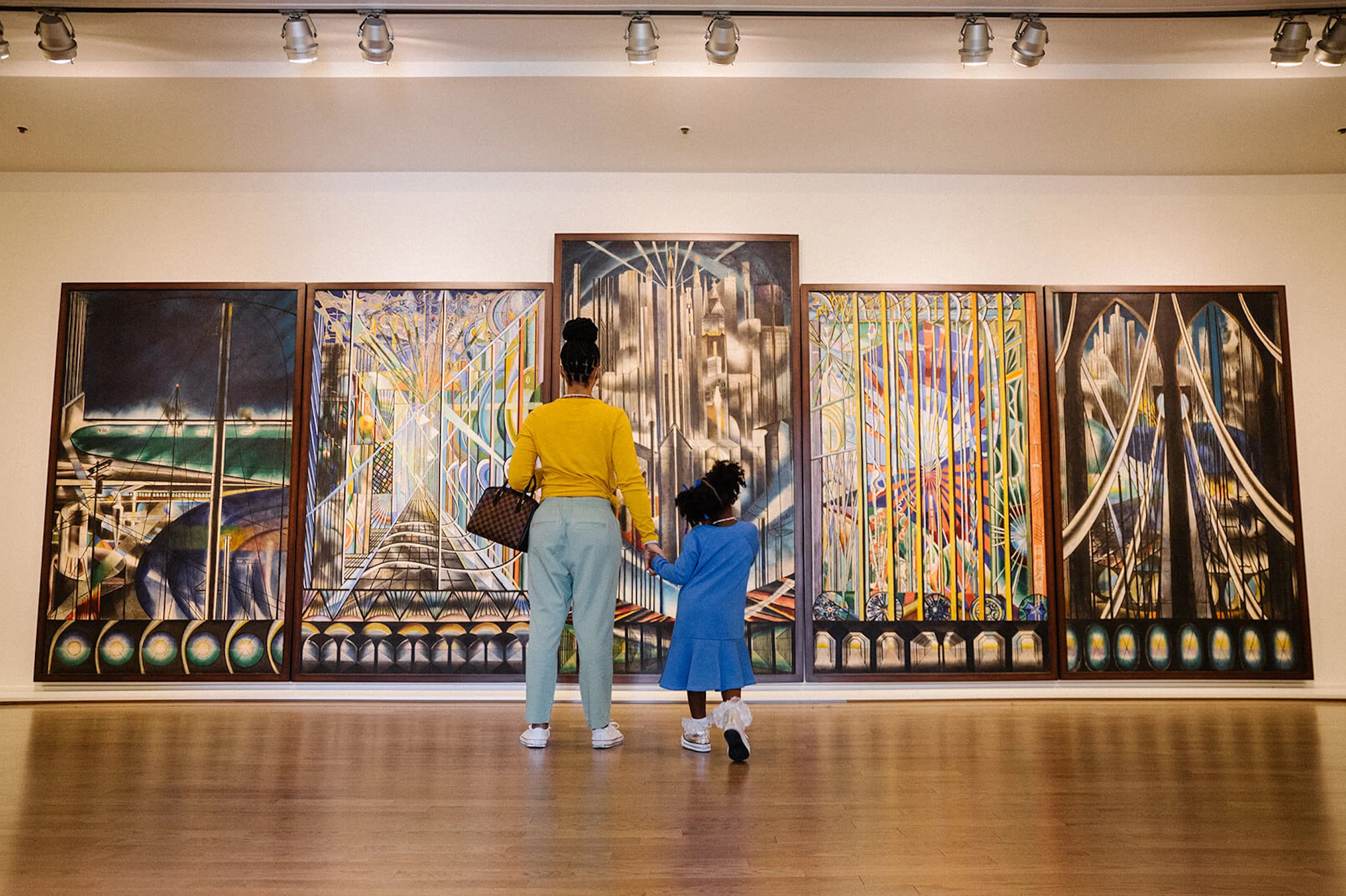 A woman and child stand before a five-paneled painting in a gallery. The panels each depict abstract scenes of bridges in fantastical colors.