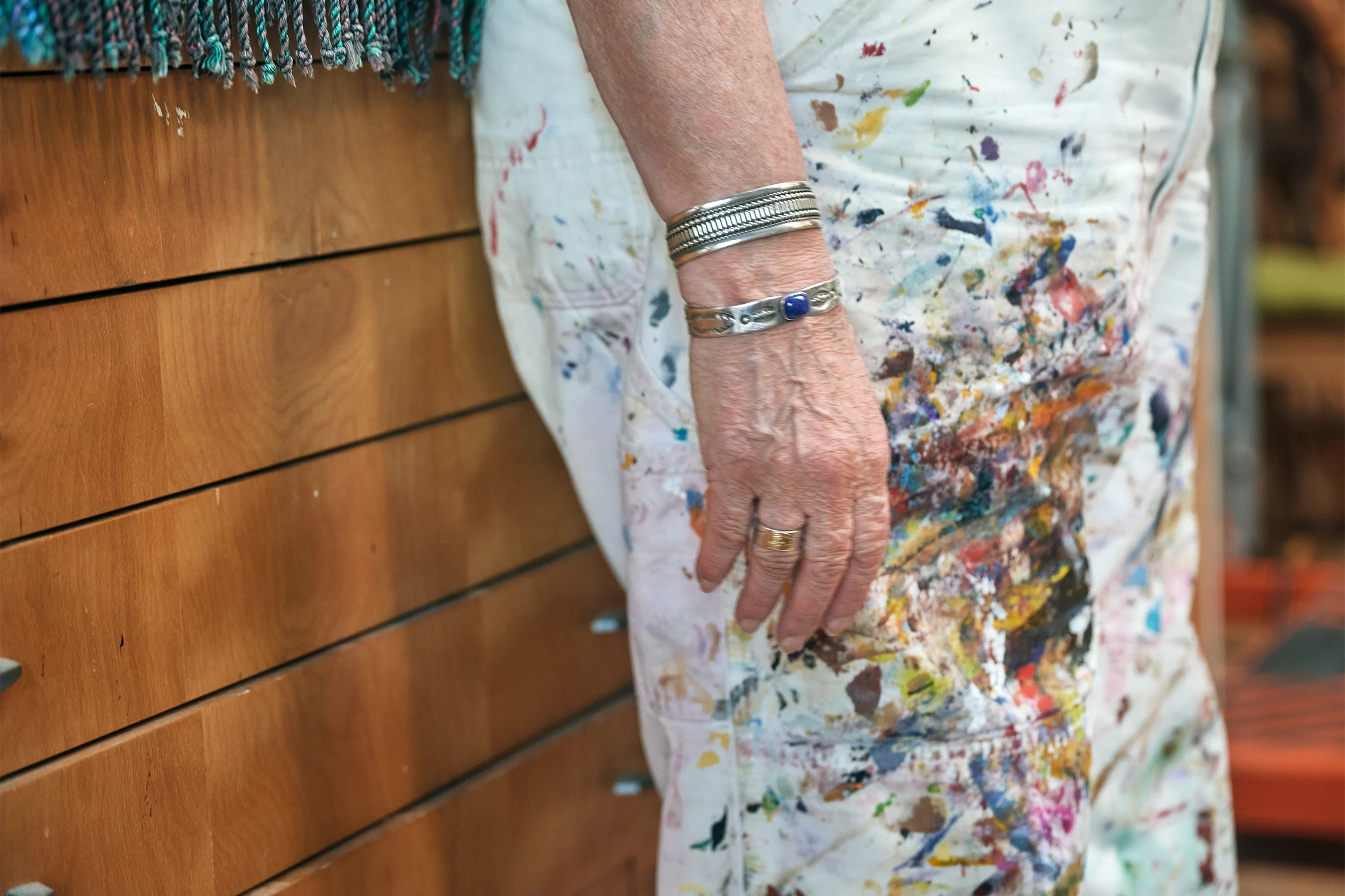 The artist Judy Baca stands in her studio wearing a white paint splattered canvas jumpsuit and metal bracelets