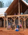 A group of Indigenous Maskoke people stand and sit in the wooden frame of a structure in a woods