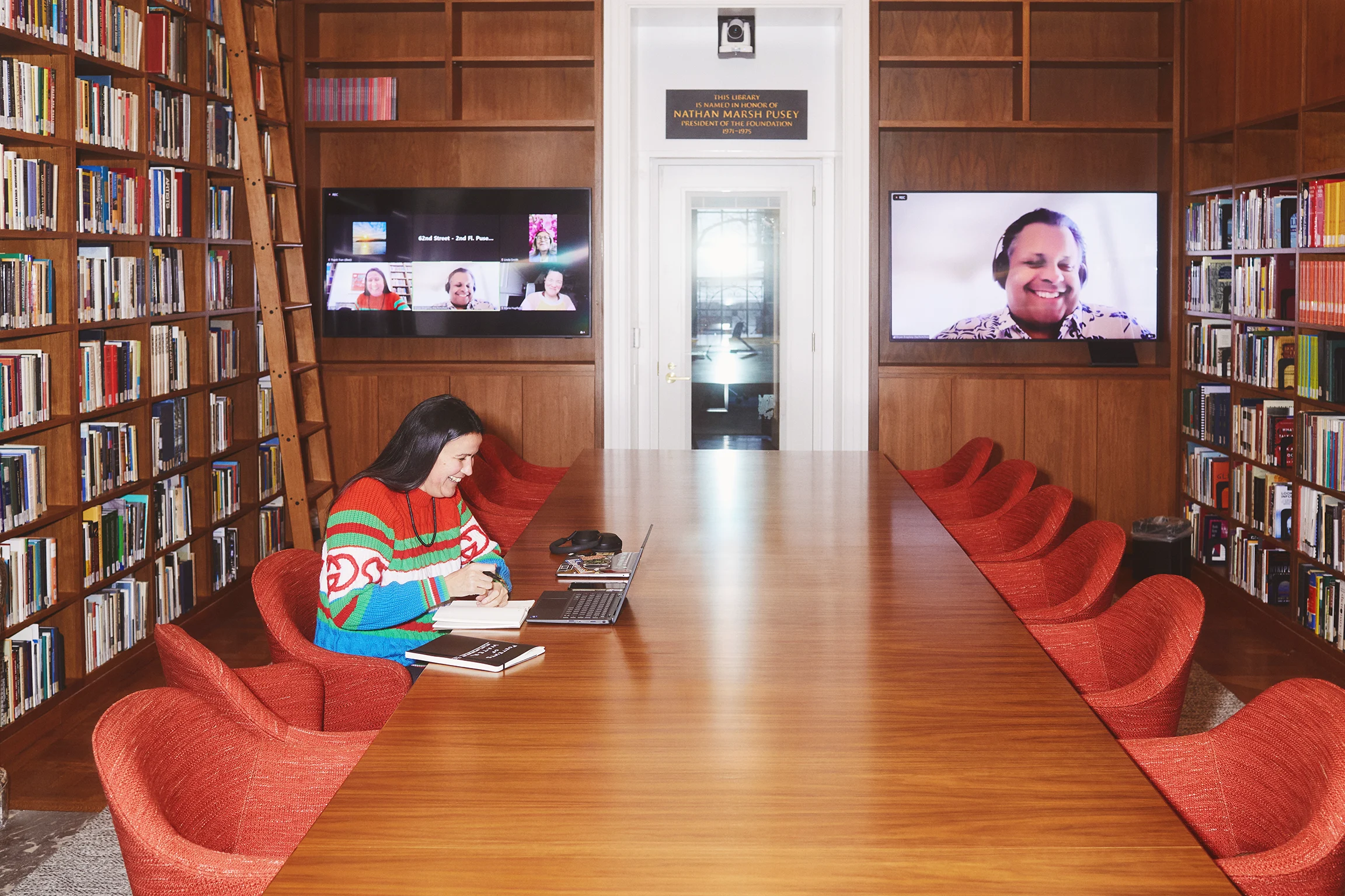 An Indigenous scholar sits at a table in a large room. Behind them are two screens with faces of people who are on a video conference call. 