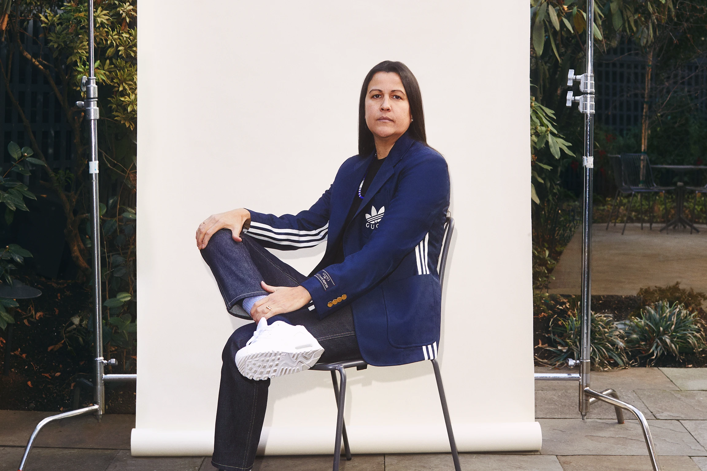 An Indigenous person wearing a sport track suit sits legs-crossed on a chair in front of a backdrop.