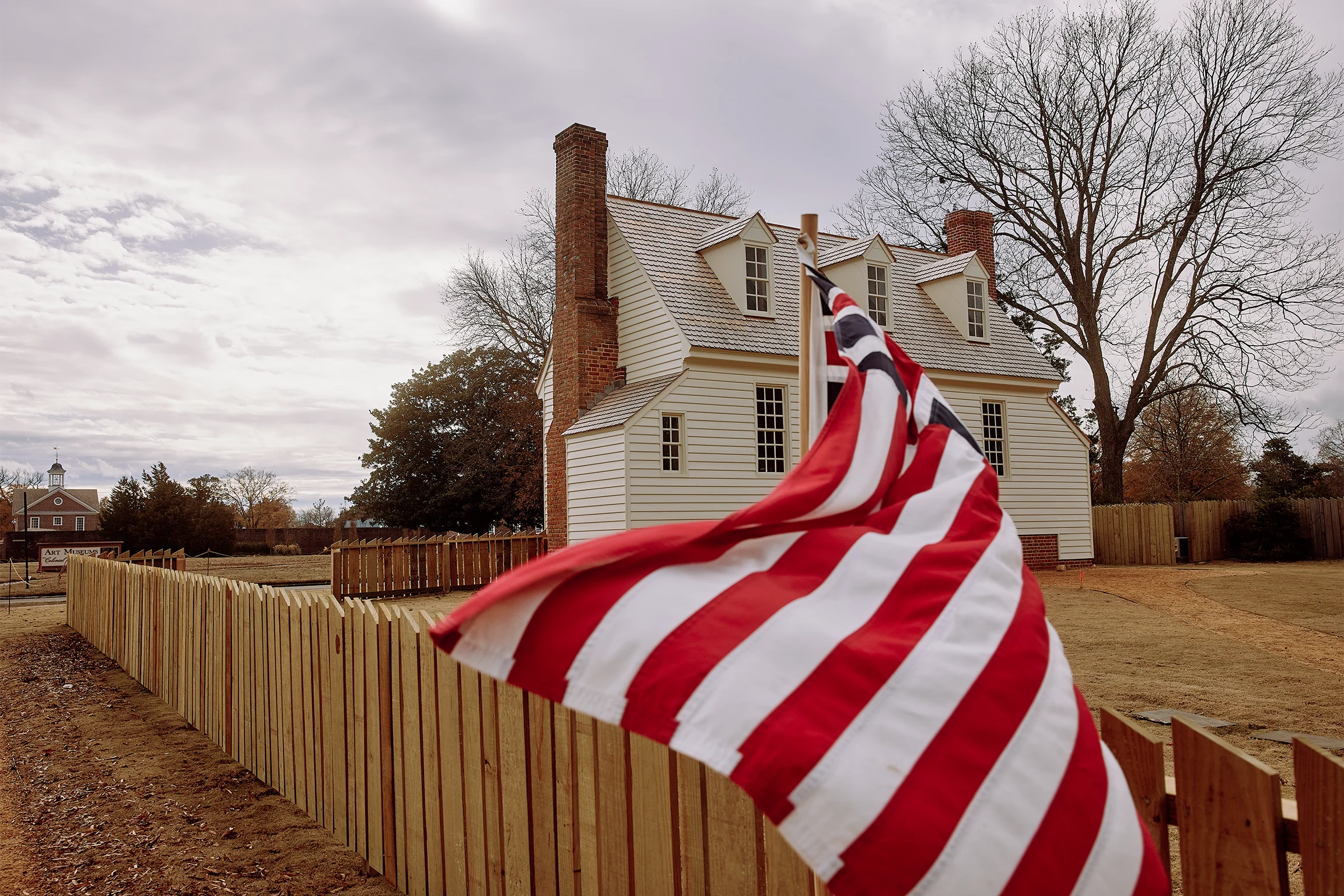 Historic two-story house with 2 chimneys, surrounded by leafless trees and a wooden fence. An American flag waves in the foreground.