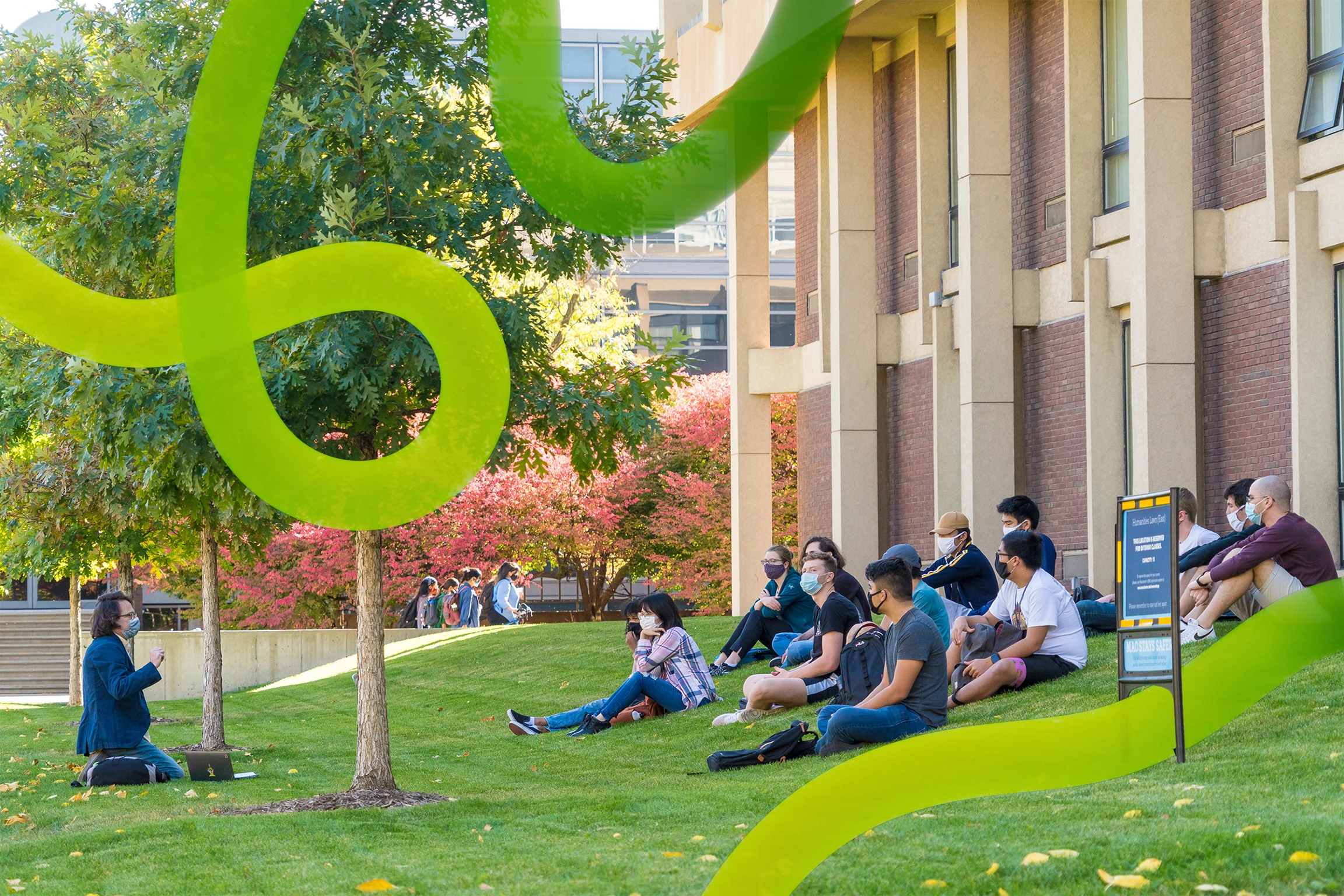 A class of college students sits on a grassy hill on a college campus. The gaze of the students is directed to an instructor, who faces the class.