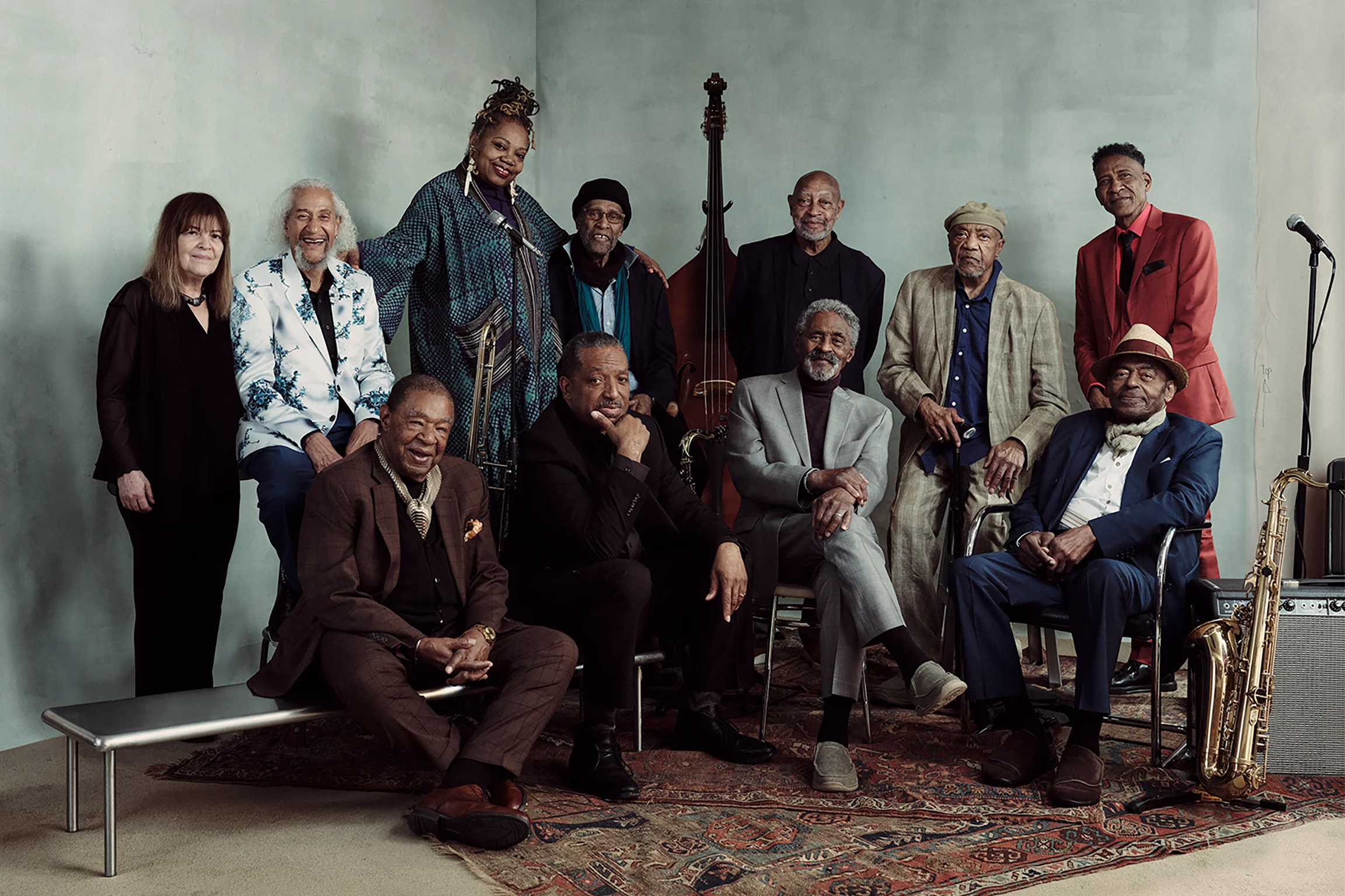 Group portrait of eleven older musicians in a studio, with some seated and others standing. A double bass stands behind them, and a saxophone rests beside an amplifier. They face the camera, smiling, against a muted blue-gray backdrop.