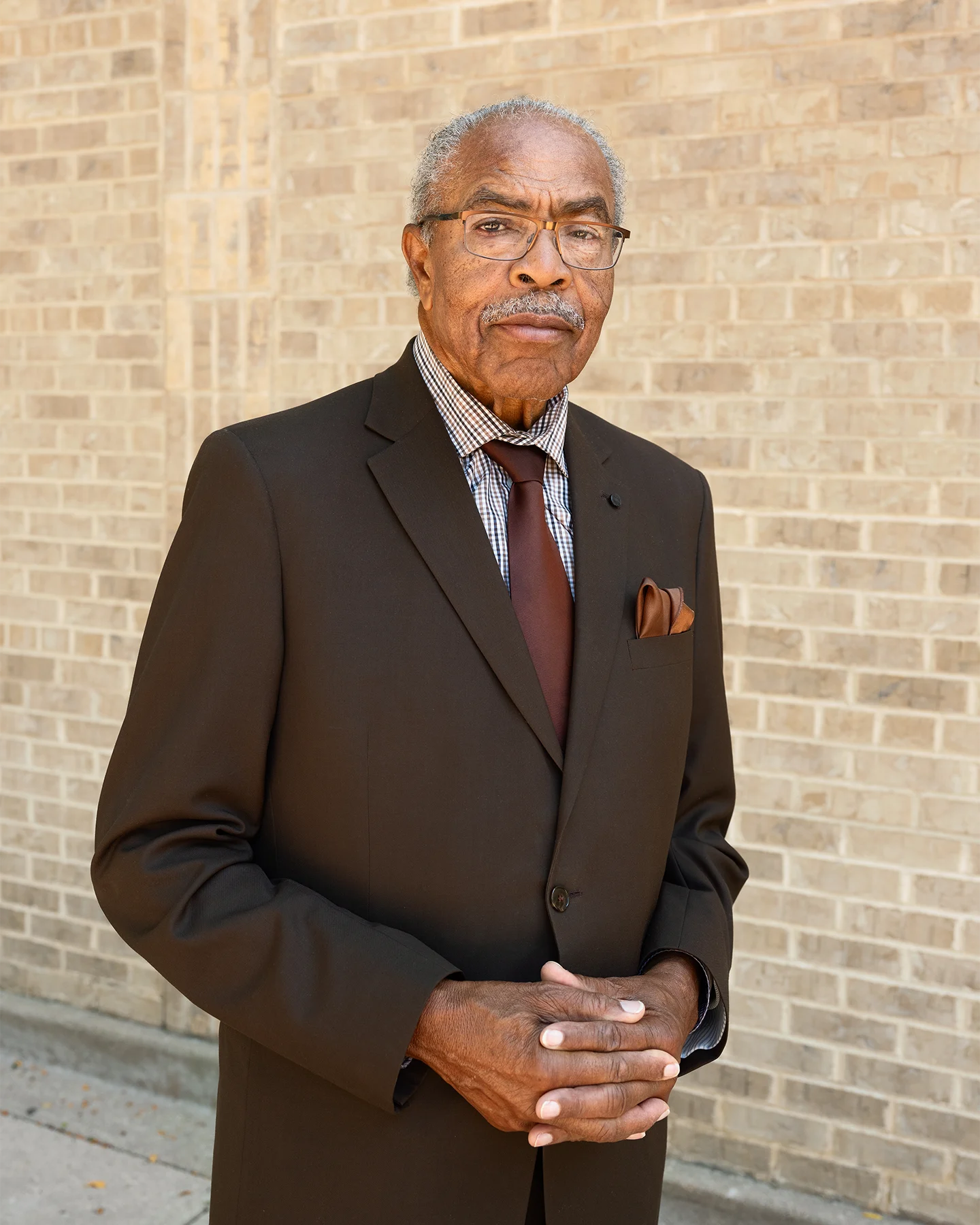 An African American man identified as Reverend Wheeler Parker Jr. standing in a dark suit and a maroon tie with glasses and his hands folded