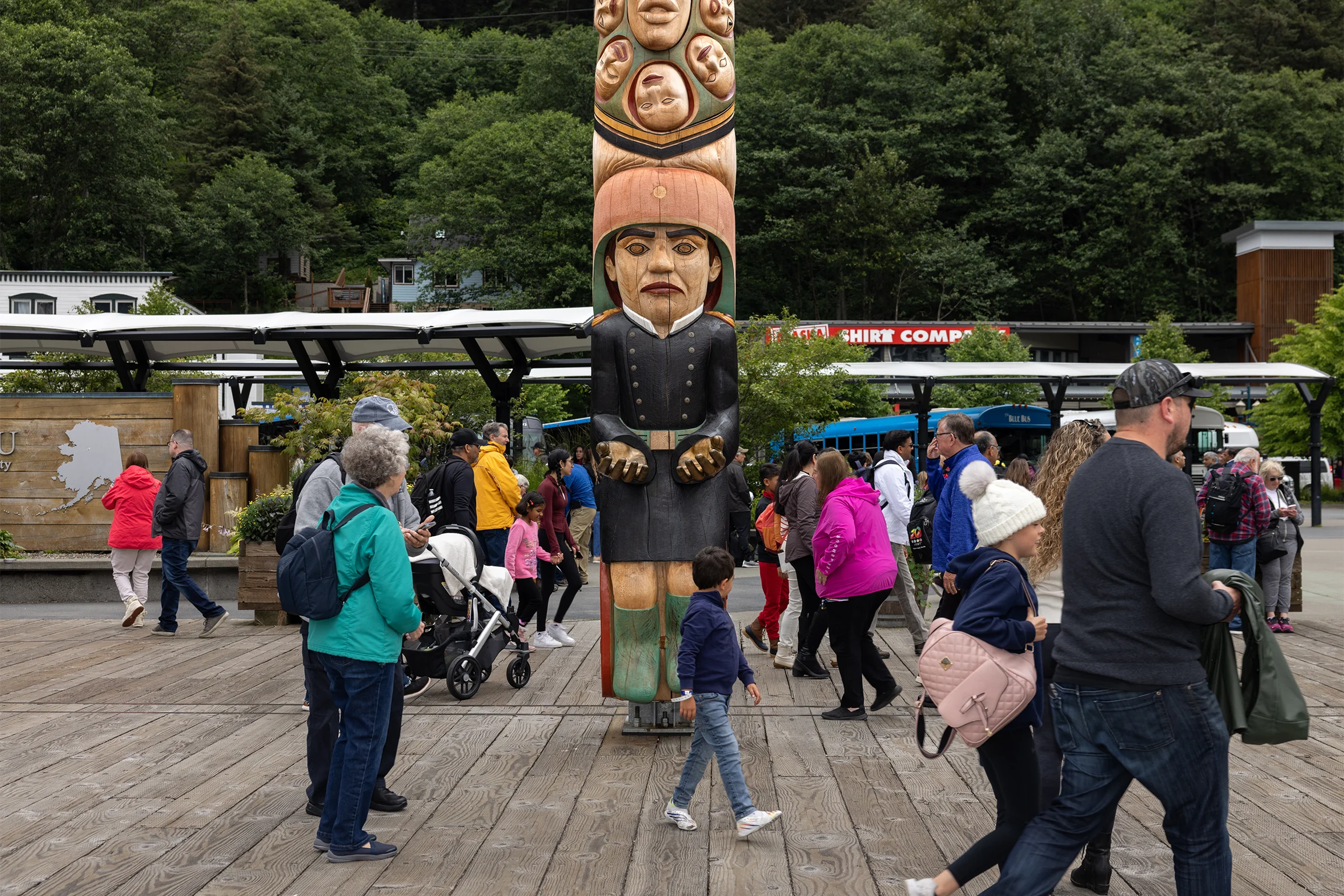A group of tourists stands on a dock looking at a large totem pole