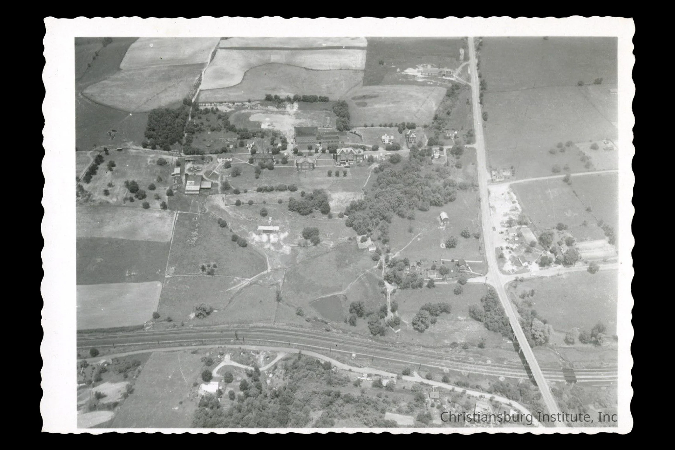 Black-and-white aerial photograph of a rural campus surrounded by farmland, roads, and railroad tracks, with several buildings and open fields visible.