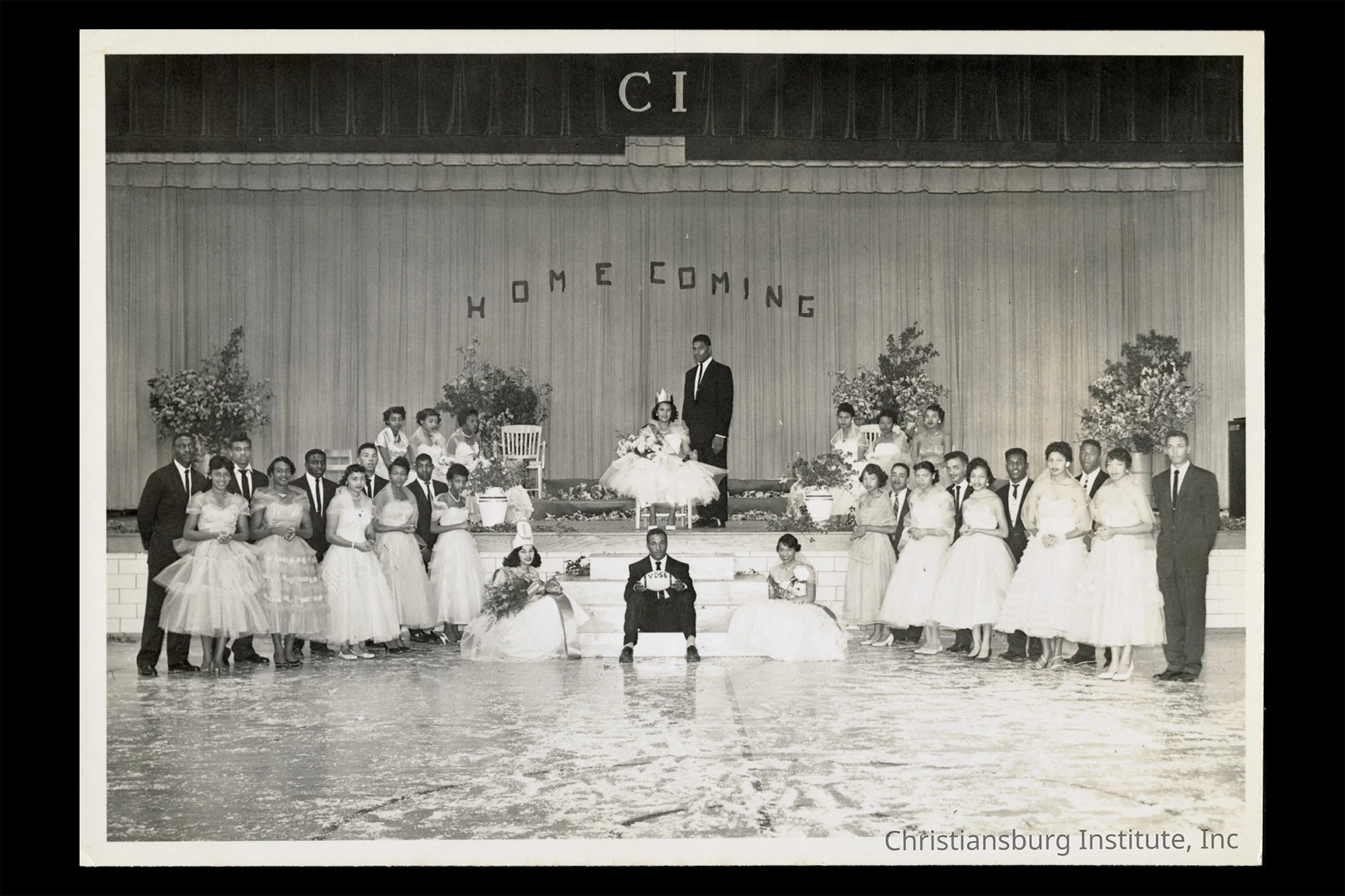 Black-and-white group portrait from a school homecoming event, showing formally dressed students gathered onstage around a seated homecoming queen beneath a “HOMECOMING” sign.
