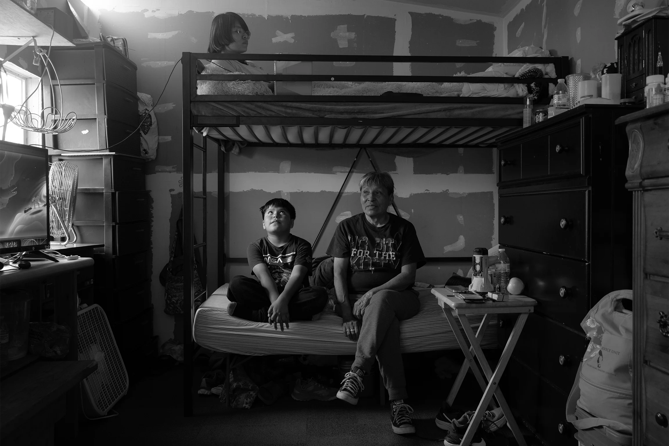 A black-and-white image shows a small, cluttered bedroom with a bunk bed. An adult and a child sit on the lower bed, looking upward, while another person lies on the top bunk. Dressers, a TV, and personal items fill the tight space.
