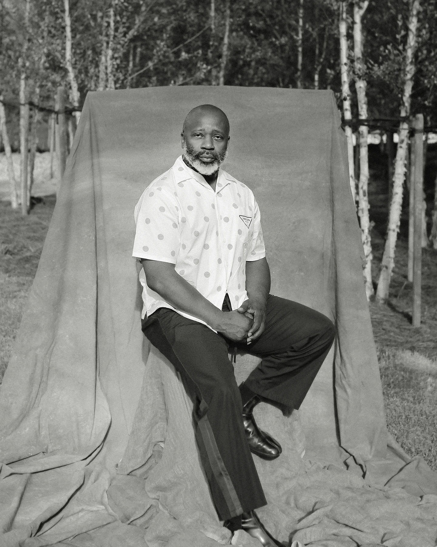 A Black man identified as the artist Theaster Gates wearing a white shirt and dark pants sits on a stool covered with a tarp