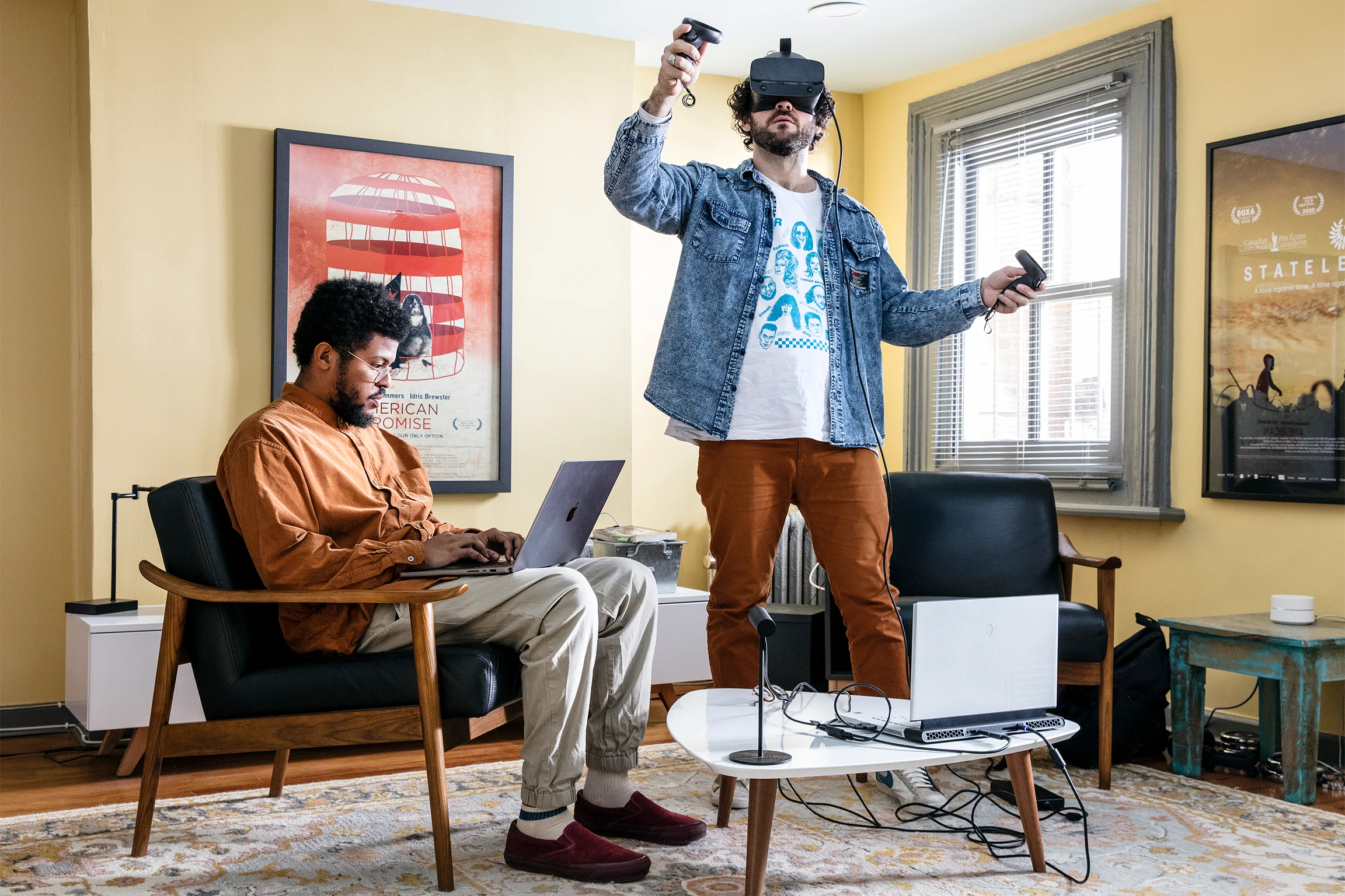A man sits in a chair with a laptop on his lap while another man stands in front of him while using a virtual reality headset