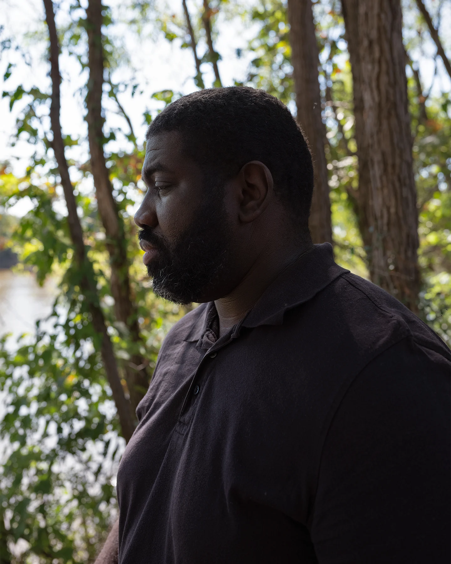 The profile of an African American man with a beard standing on the side of a river with greenery behind him