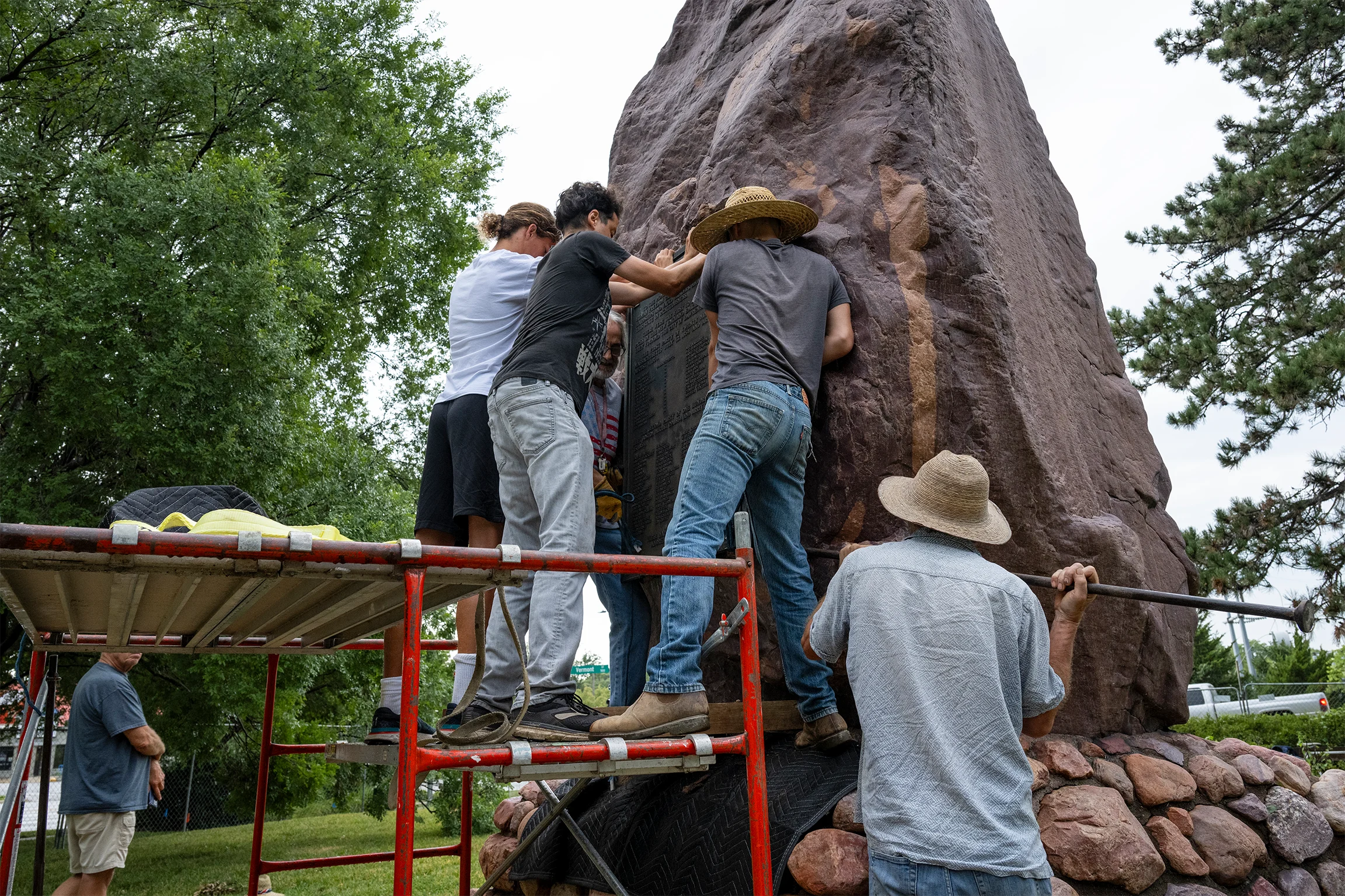 A group of workers stands on steel scaffolding while removing a plague from a large rock